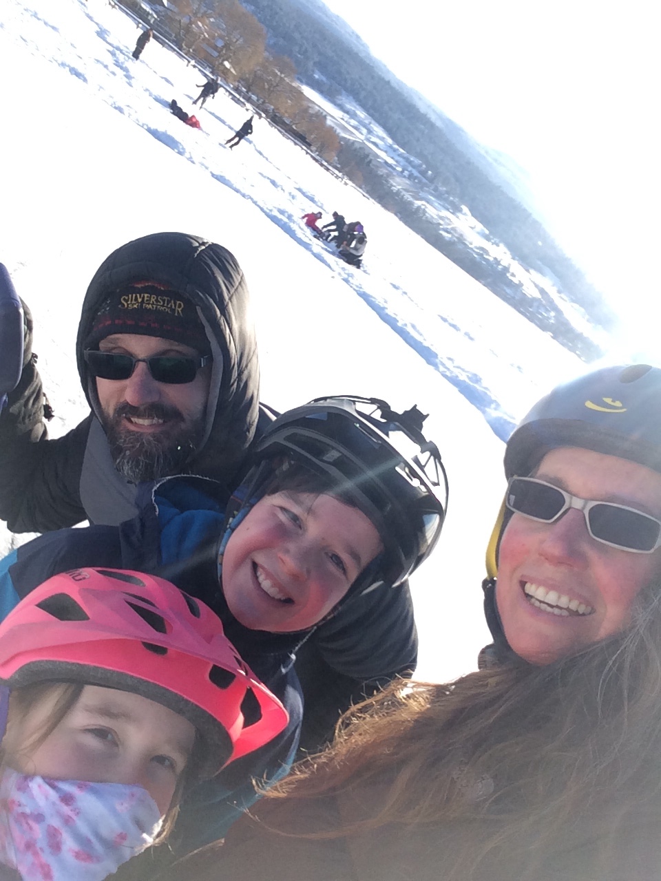 Family with mom and dad wearing sunglasses; father wearing hood with beard and moustache; mother, boy and girl wearing helmets; skating rink and mountain in background
