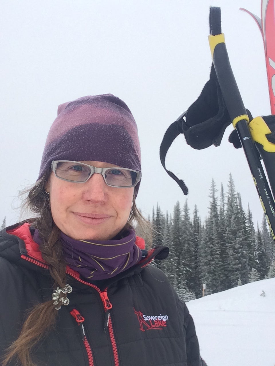 Woman with long hair in a braid, wearing hat, glasses and jacket, holding skis and poles with snow and evergreen forest in background