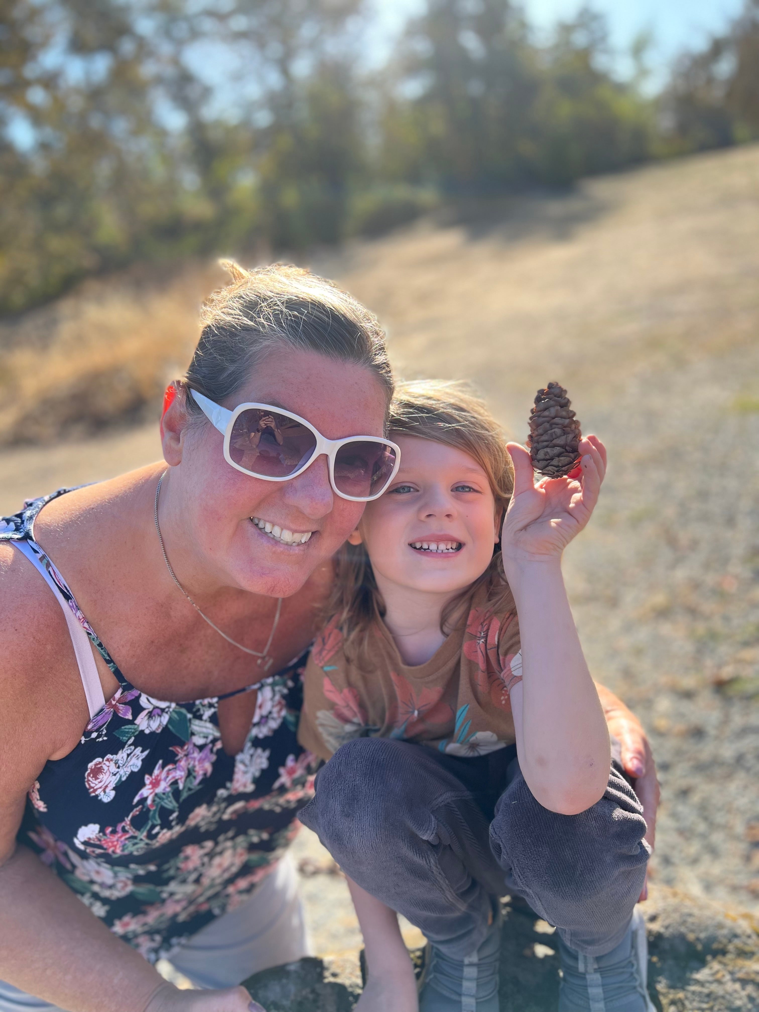 A woman and a child holding a pinecone in front of a forest 