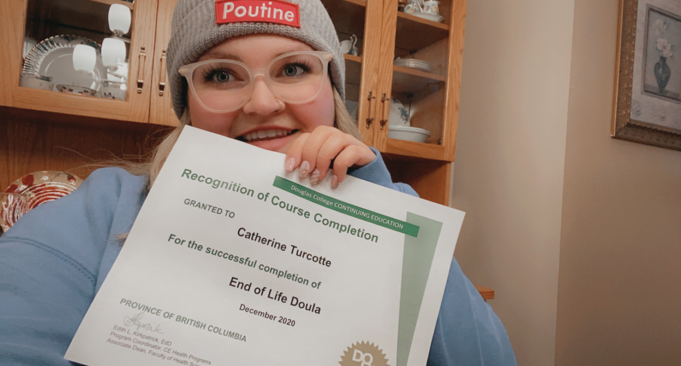 Smiling woman wearing grey poutine toque and glasses, holds End of Life Doula certificate