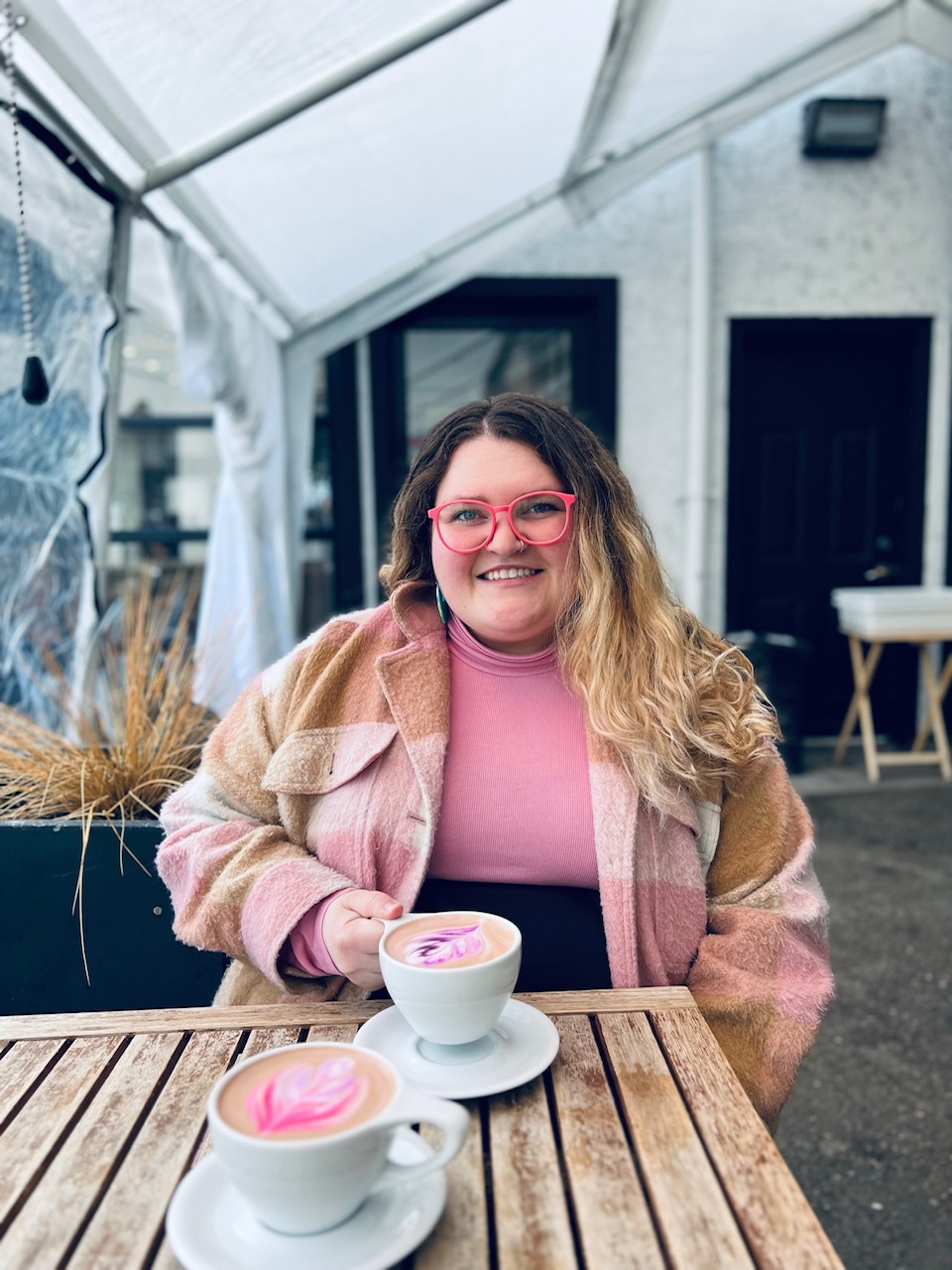 Smiling woman with long hair and glasses, seated at wooden table, wearing pink top and jacket, with cup of hot chocolate
