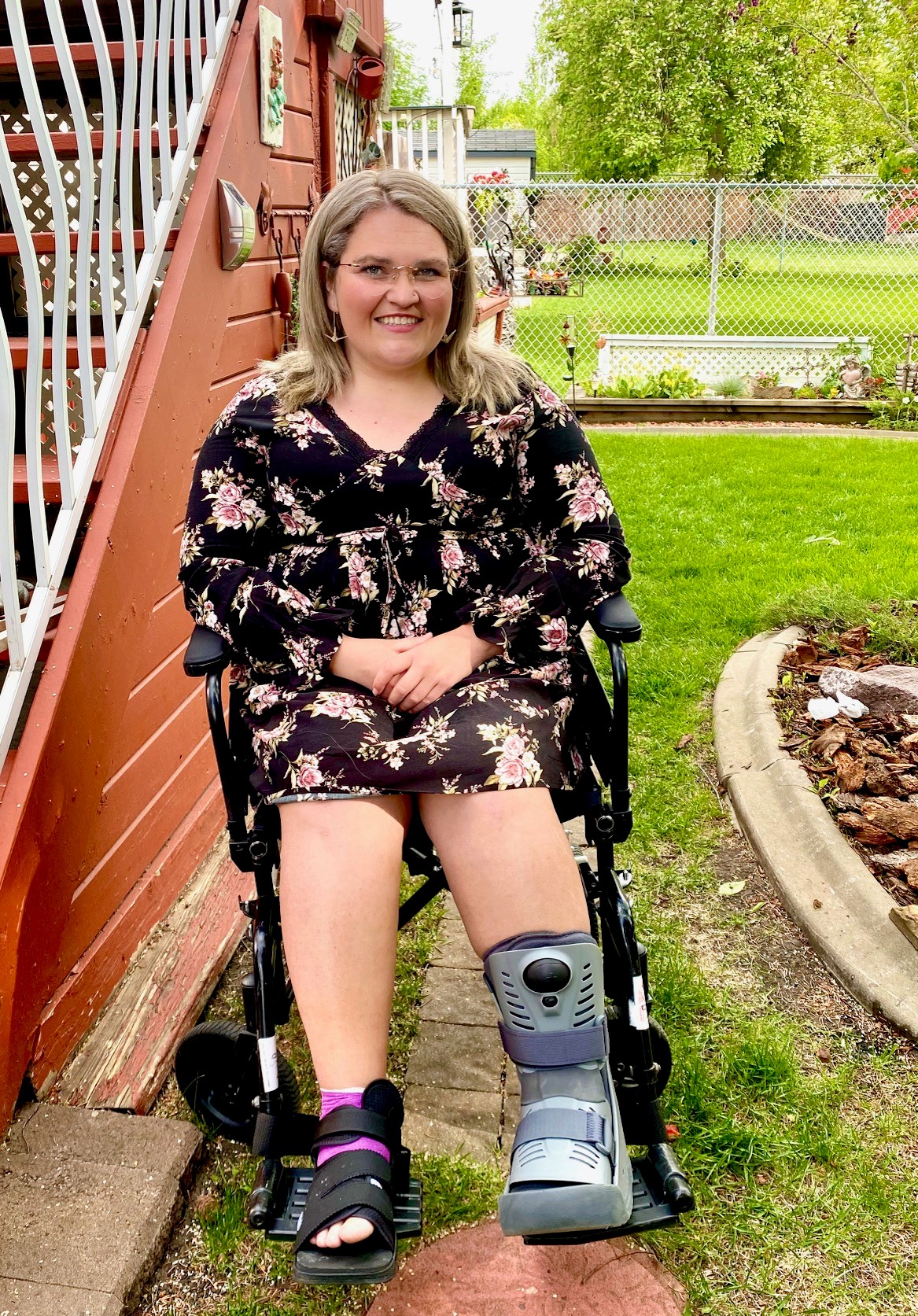 Smiling woman with long blonde hair wearing glasses, long-sleeved black patterned dress, foot brace and ankle brace, sits in wheelchair on grass lawn in backyard next to a flight of stairs with white handrail