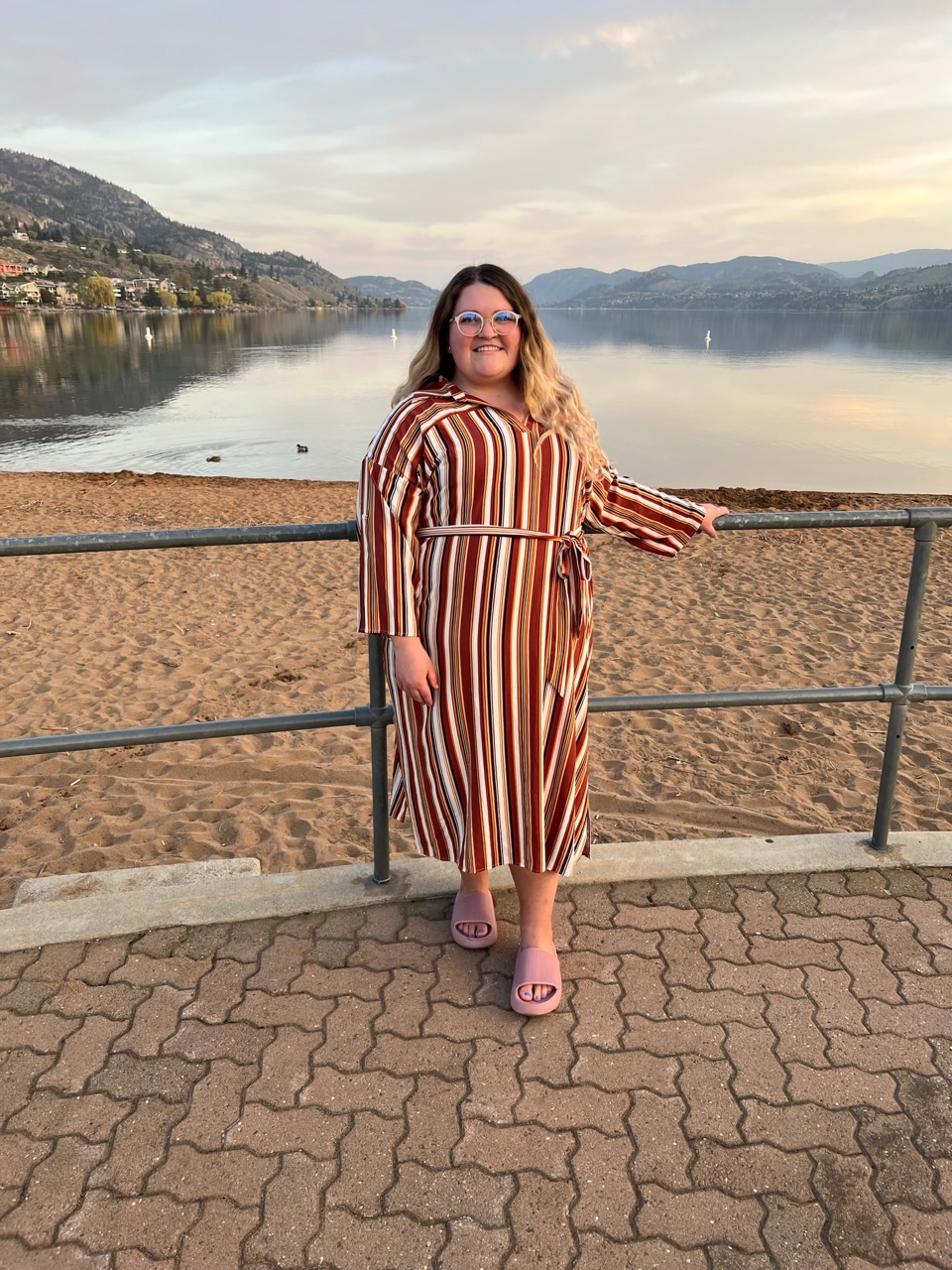 Smiling woman with long hair and glasses, wearing long striped dress with belt, sliders sandals, standing leaning against rail by beach at lake with mountains in the distance