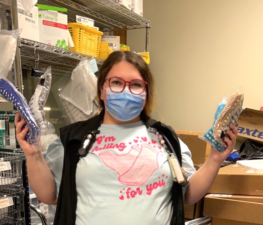 Woman in storage supply room with blue face mask and glasses on, wearing t-shirt with socks and holding non-slip socks