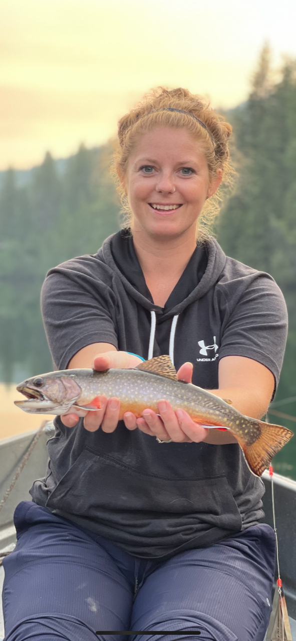 Woman holding fish by lake and trees.