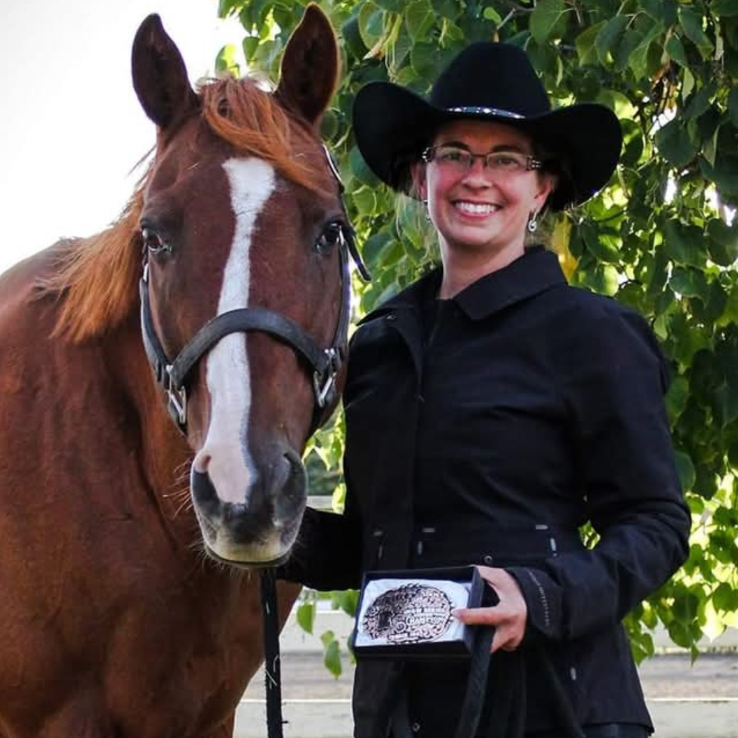  A person wearing glasses and a cowboy hat stands next to a brown horse with a white stripe on its nose. The person holds an award. 