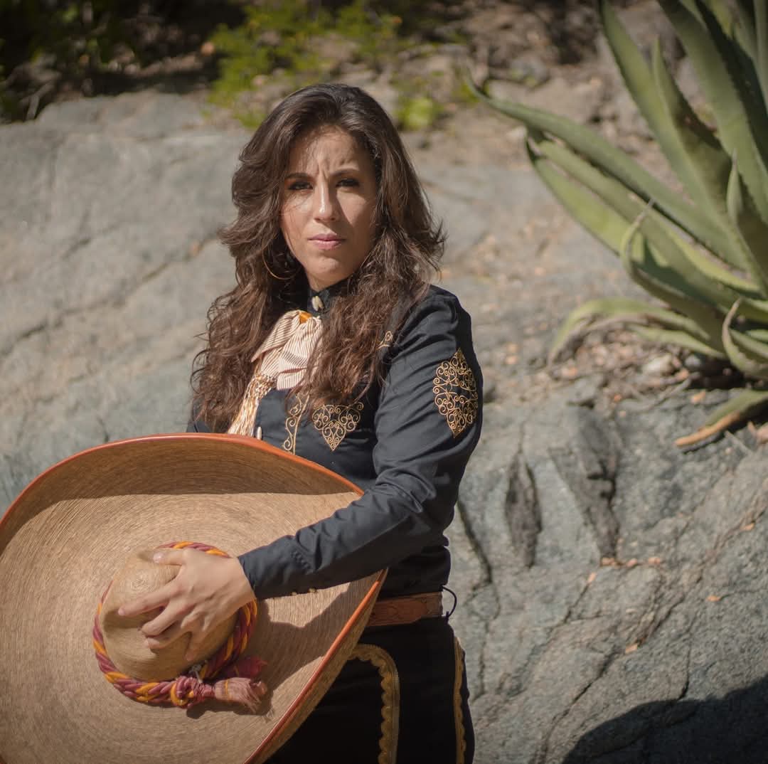 A Mexican woman with long dark curled hair is wearing a black traditional, black with rose gold/orange embroidery, Mexican, charro de feana suit. She is holding a sombrero with one hand over her abdomen looking at the camera.
