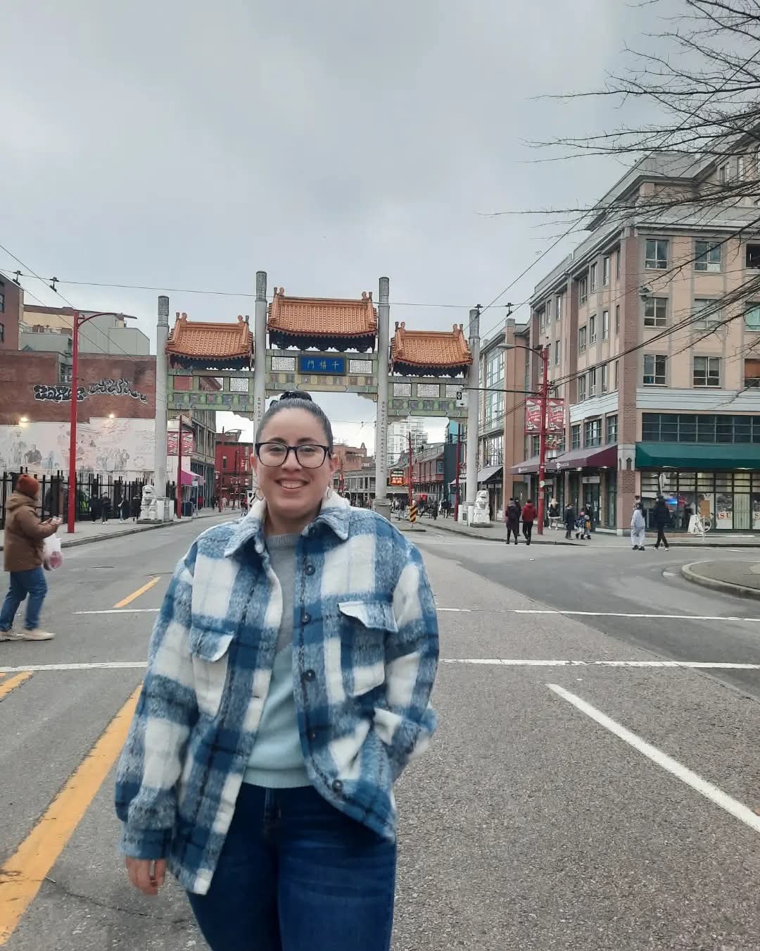 A woman with dark hair pulled back into a bun wearing dark rimmed glasses, dark blue jeans and a blue and white plaid shacket is standing with one hand in her pocket smiling at the camera. She is in the middle of a busy street in China town in Vancouver B.C. 