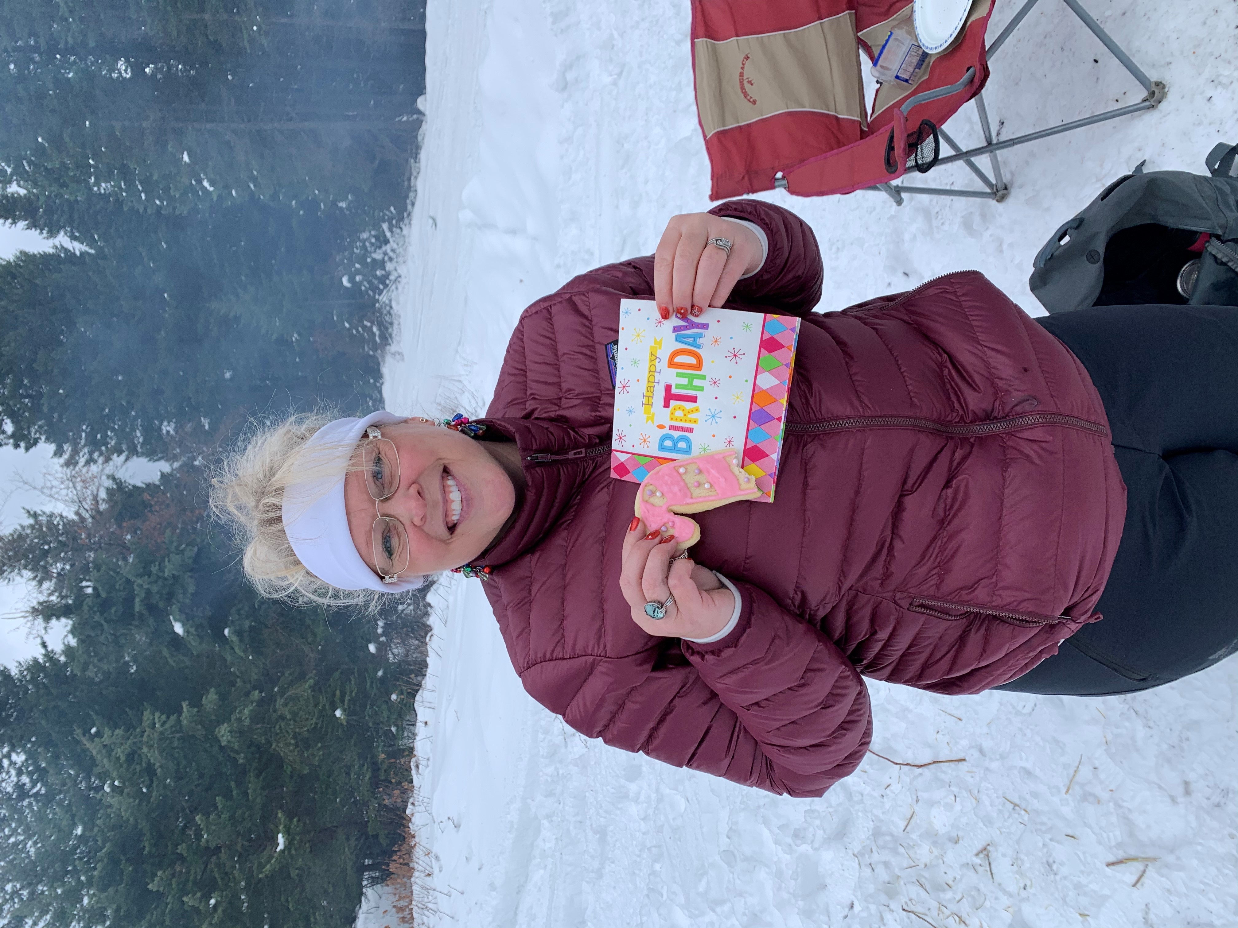 Outdoors, in the snow, a smiling woman wearing glasses, a burgundy ski jacket and a white headband, holds up a birthday card.