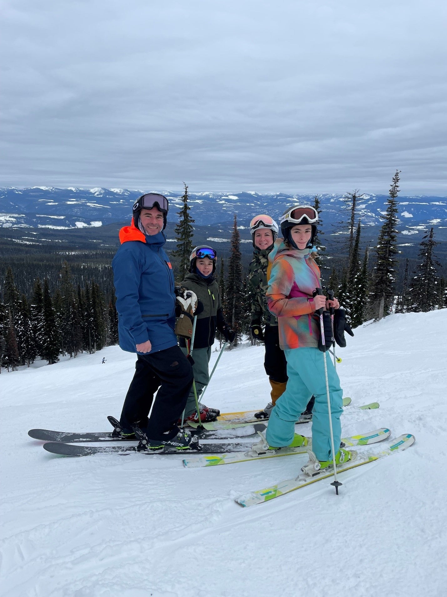 Woman, man, girl, and boy in winter clothing and skis on a snowy slope with trees in the background