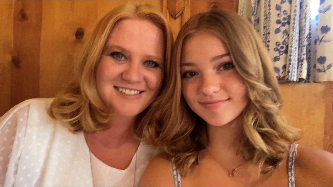 Two women, mother and daughter, with long blonde hair smile with wood panel wall and white and blue flower pattern curtain in background