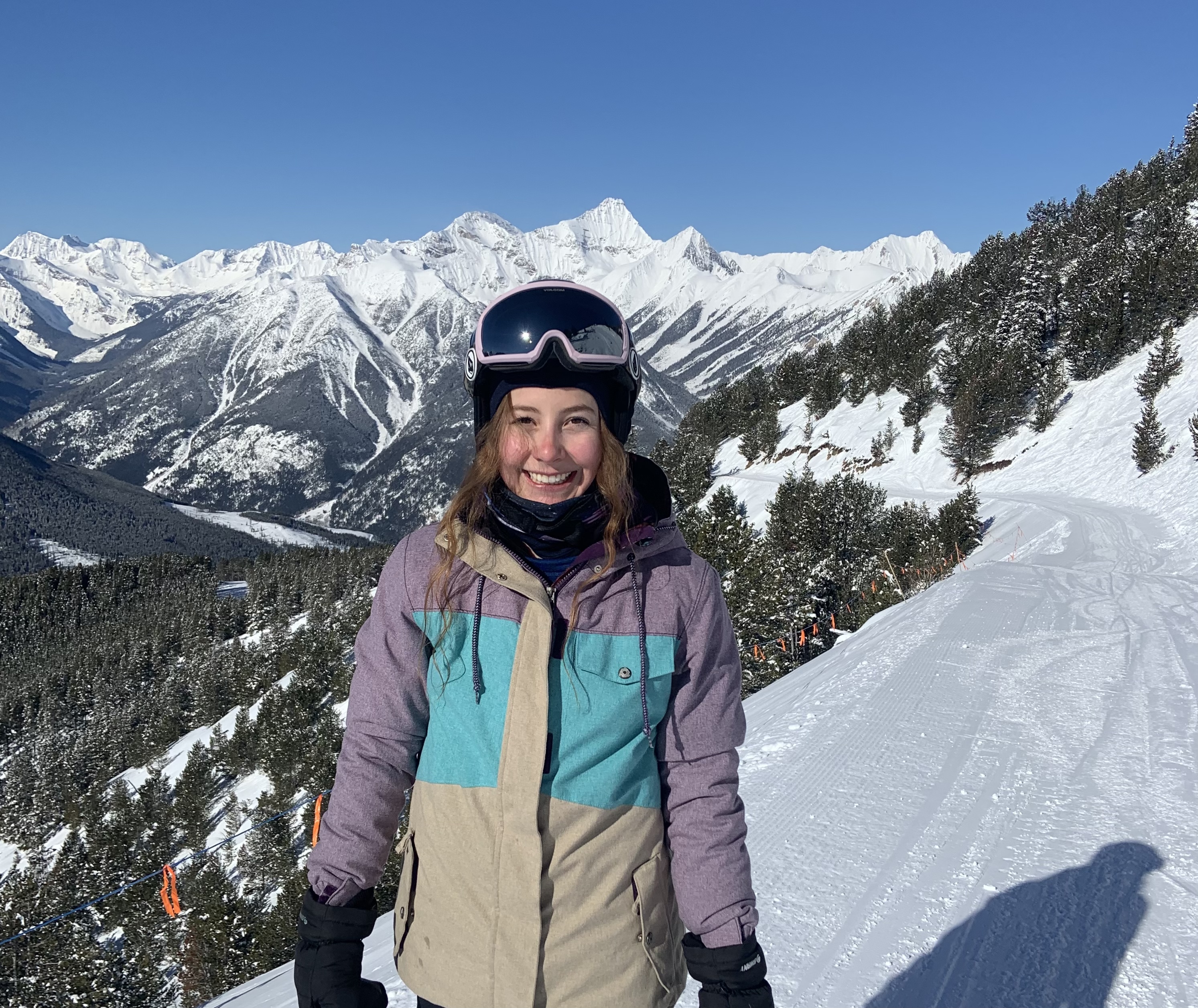 A smiling woman with long brown hair in a purple, grey and aqua winter jacket and ski goggles standing on a snowy mountain.