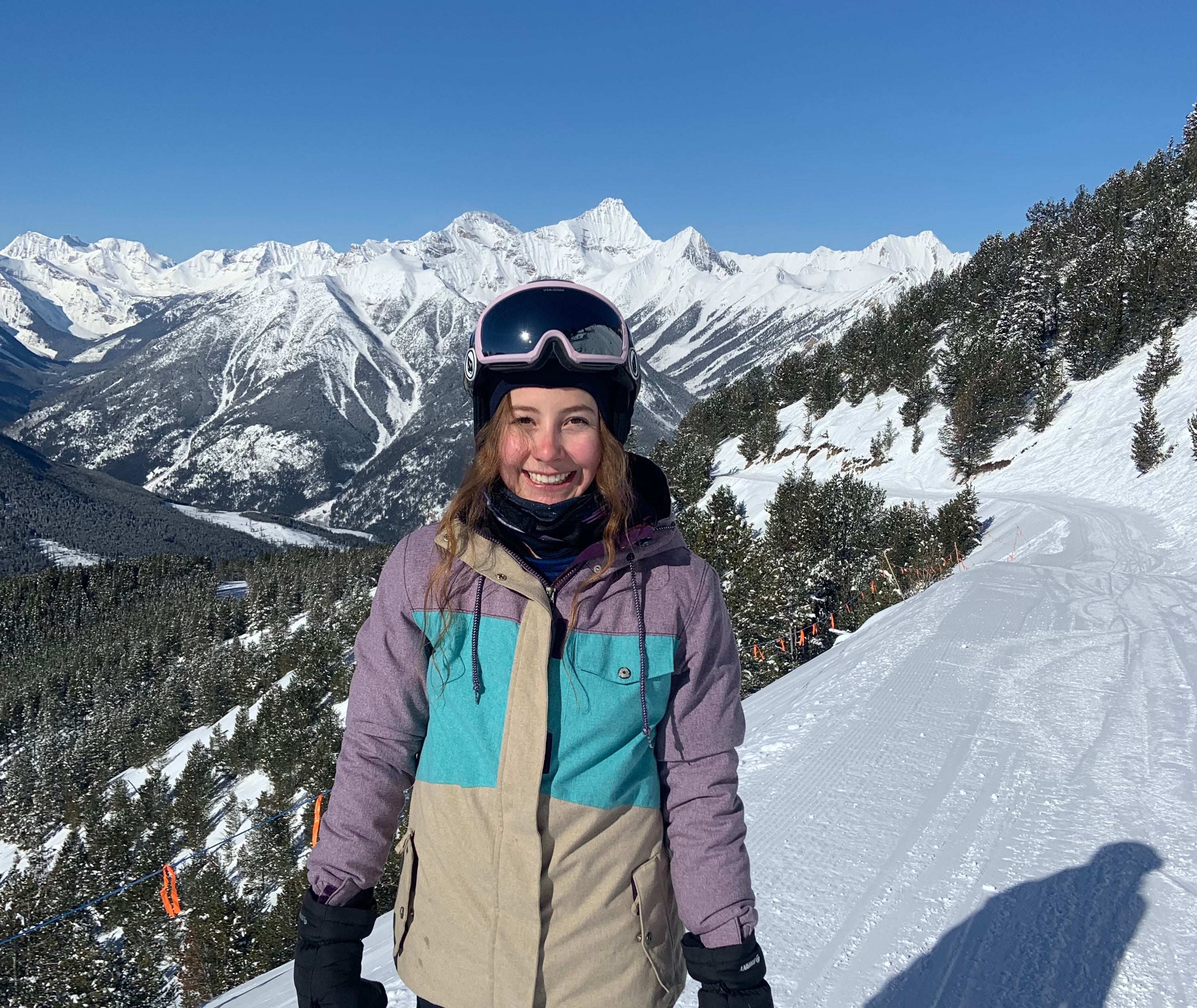 A smiling woman with long brown hair in a purple, grey and aqua winter jacket and ski goggles standing on a snowy mountain.