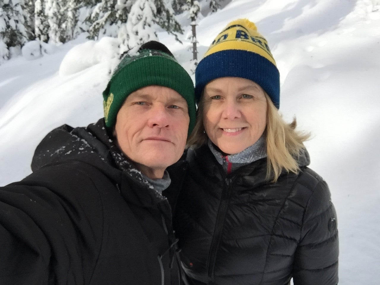 A selfie of a man on the left, wearing a green toque and black jacket, and a woman on the right, wearing a yellow and blue toque and black jacket, standing in front of snow and snowy trees.