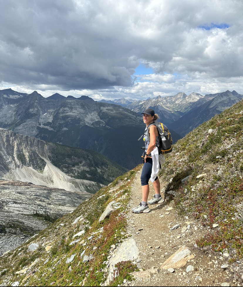 Woman with grey ballcap, sleeveless top, knee-length black pants, running shoes and backpack standing on narrow path on the side of a mountain overlooking rocky mountains and cloudy sky in the background