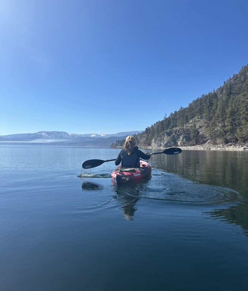 Woman kayaking in red kayak with paddle on still lake with trees, mountains and clear blue sky in background