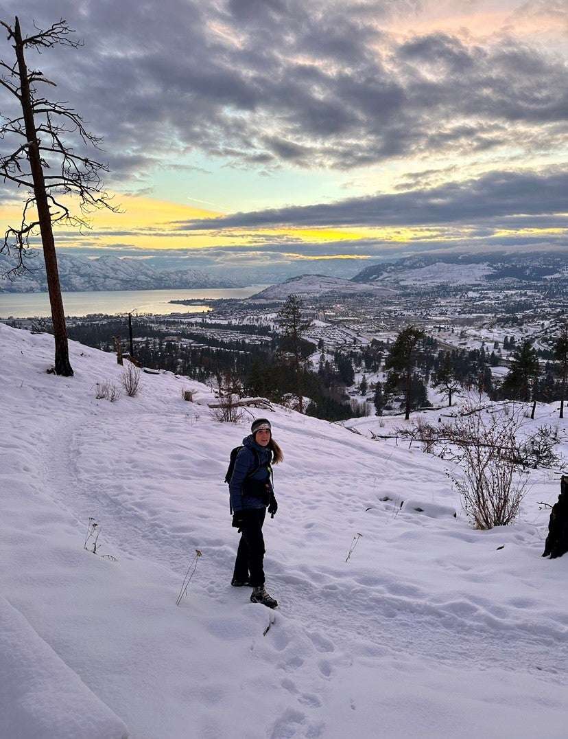 Woman with long brown hair, hat, winter jacket, black pants and boots, hiking in the snow on the side of a mountain overlooking city, trees and lake with mountains, clouds and sunset in the background