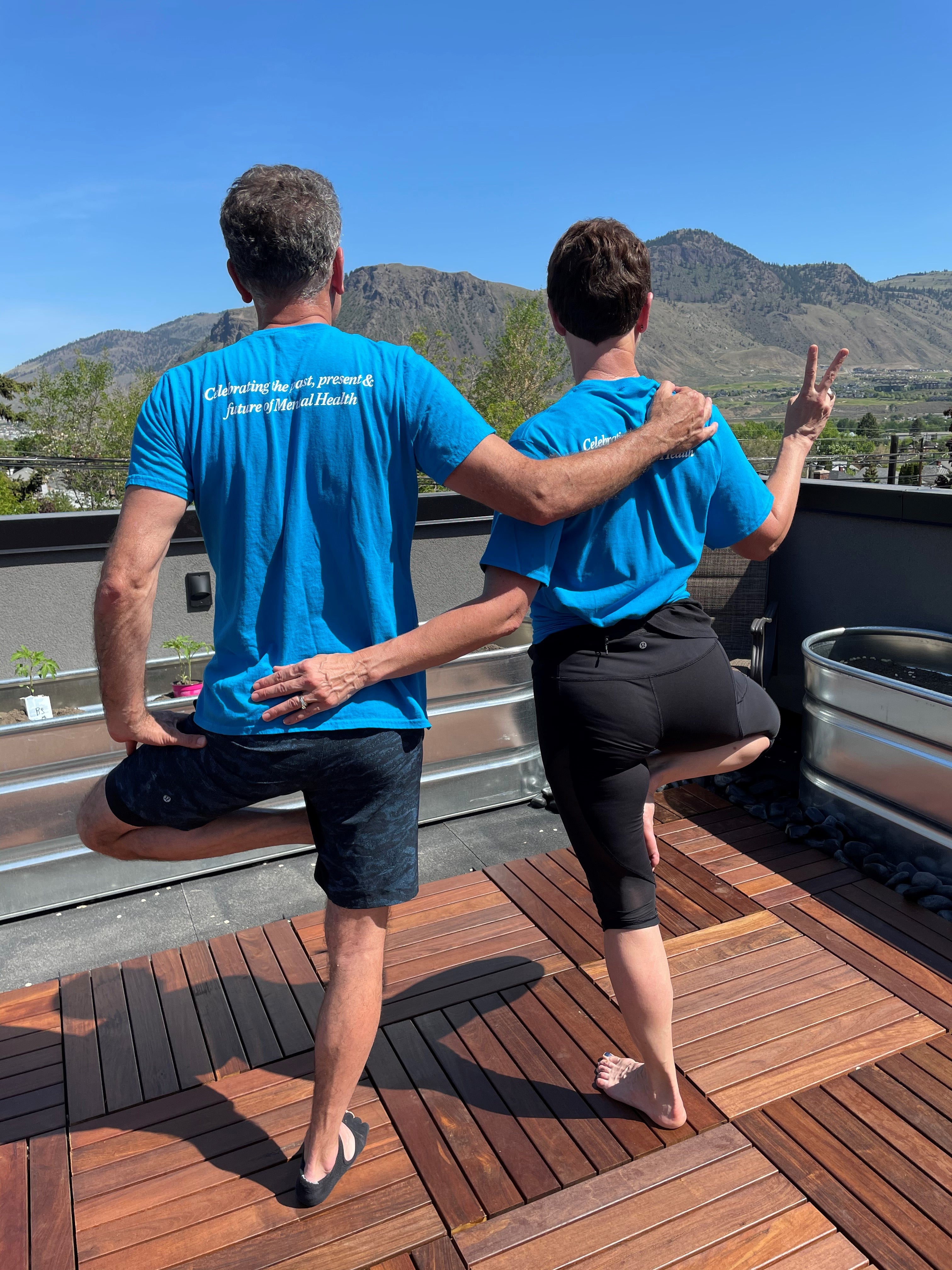 A man (left) and woman in blue shirts and yoga pants standing on one leg with the other leg at a right-angle from the standing leg, with their backs to the camera on a balcony with mountains in the background.