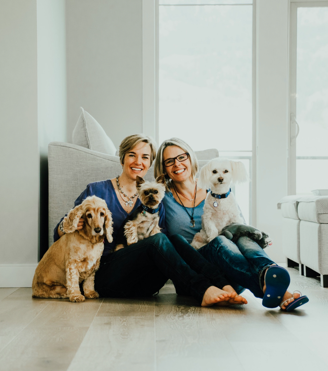 Two women with short blonde hair, smiling, wearing blue tops and jeans, with three dogs: a Spaniel, Yorkie and Maltese.