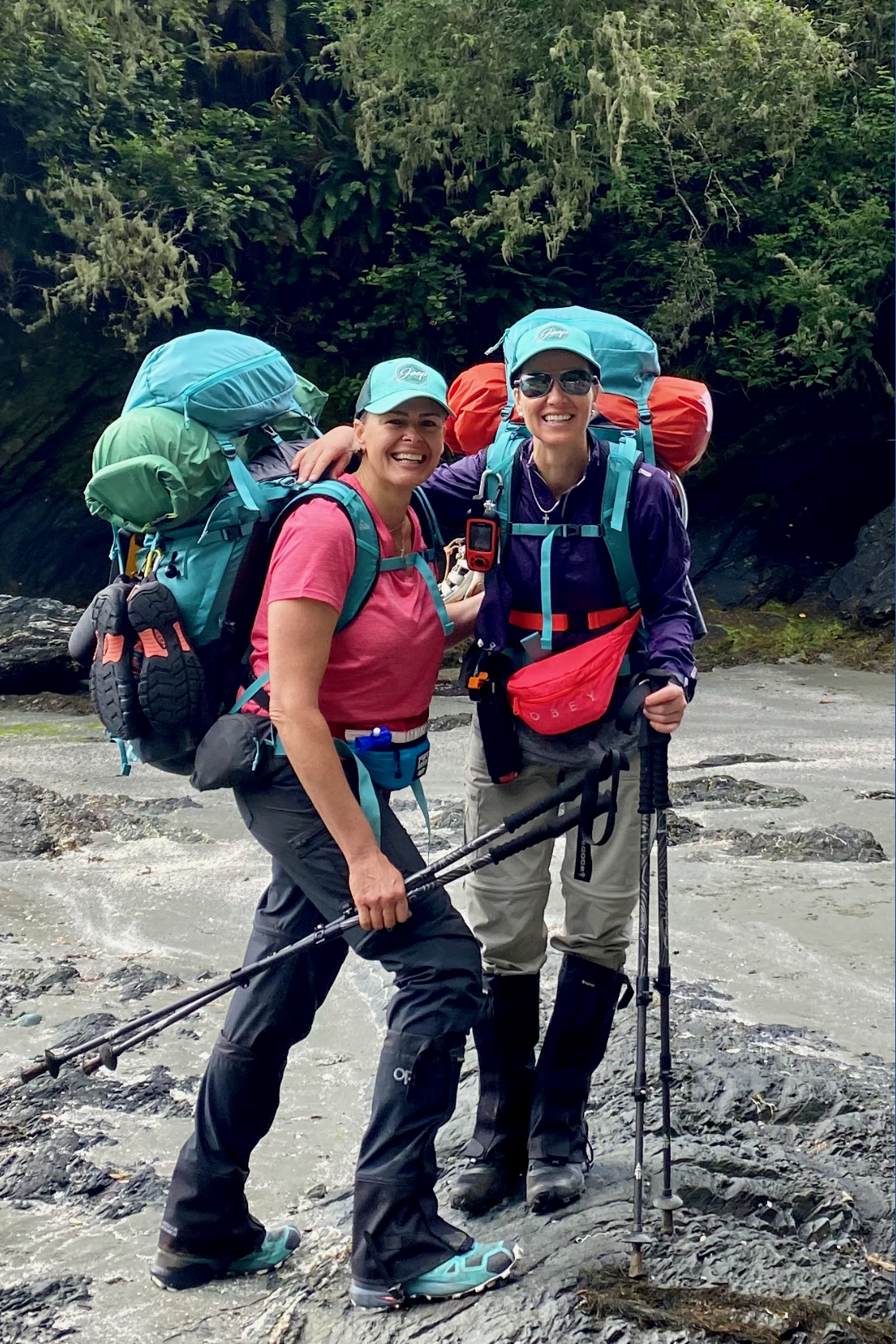 Two women wearing blue ball caps, carrying hiking polls and backpacks, with trees in the background.