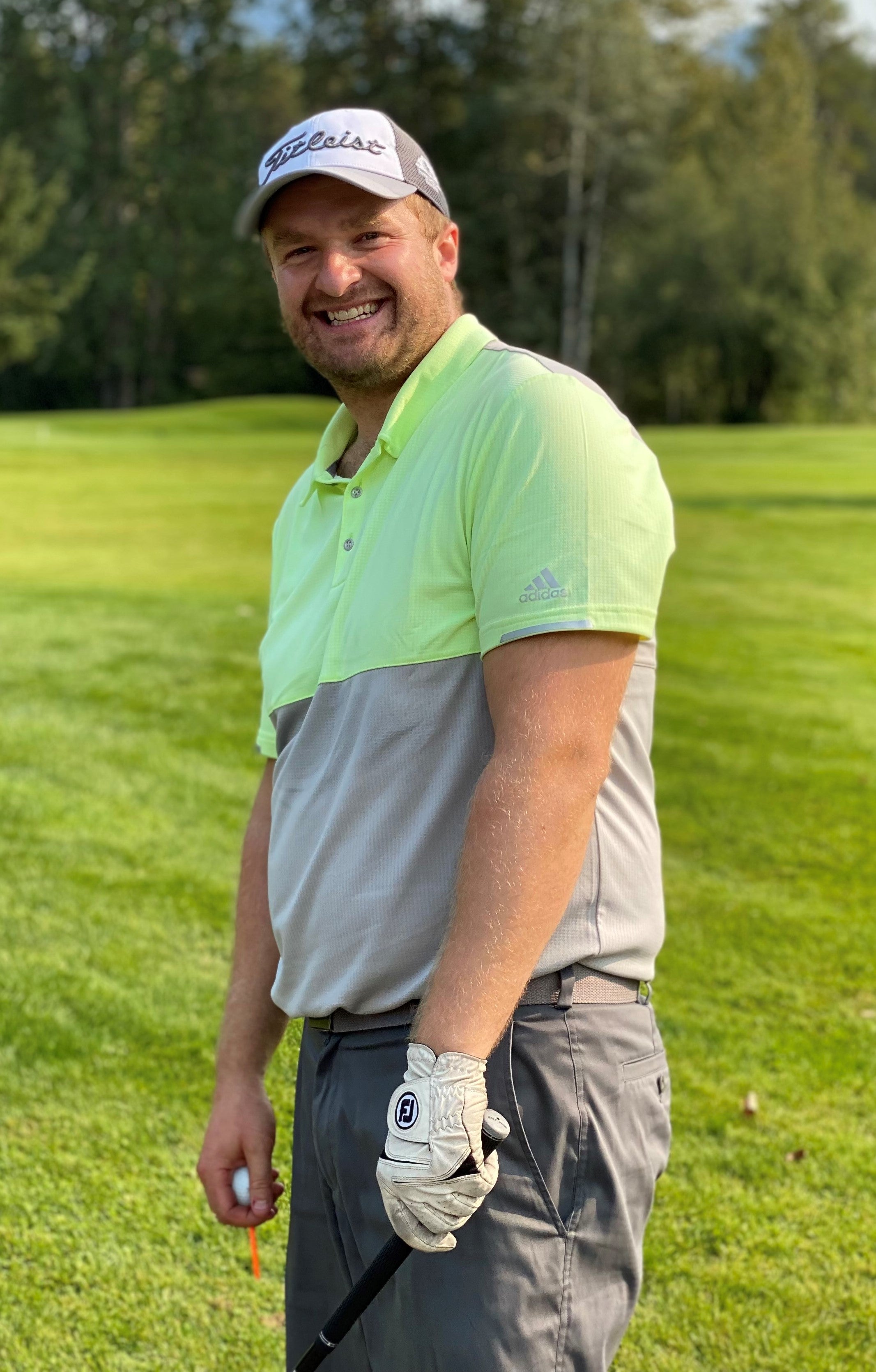 A man in a green and grey golf shirt with a grey and white ballcap, wearing a white golf glove on his left hand and holding a club while holding a white golf ball and orange golf tee in his right hand, standing in front of grass and trees.