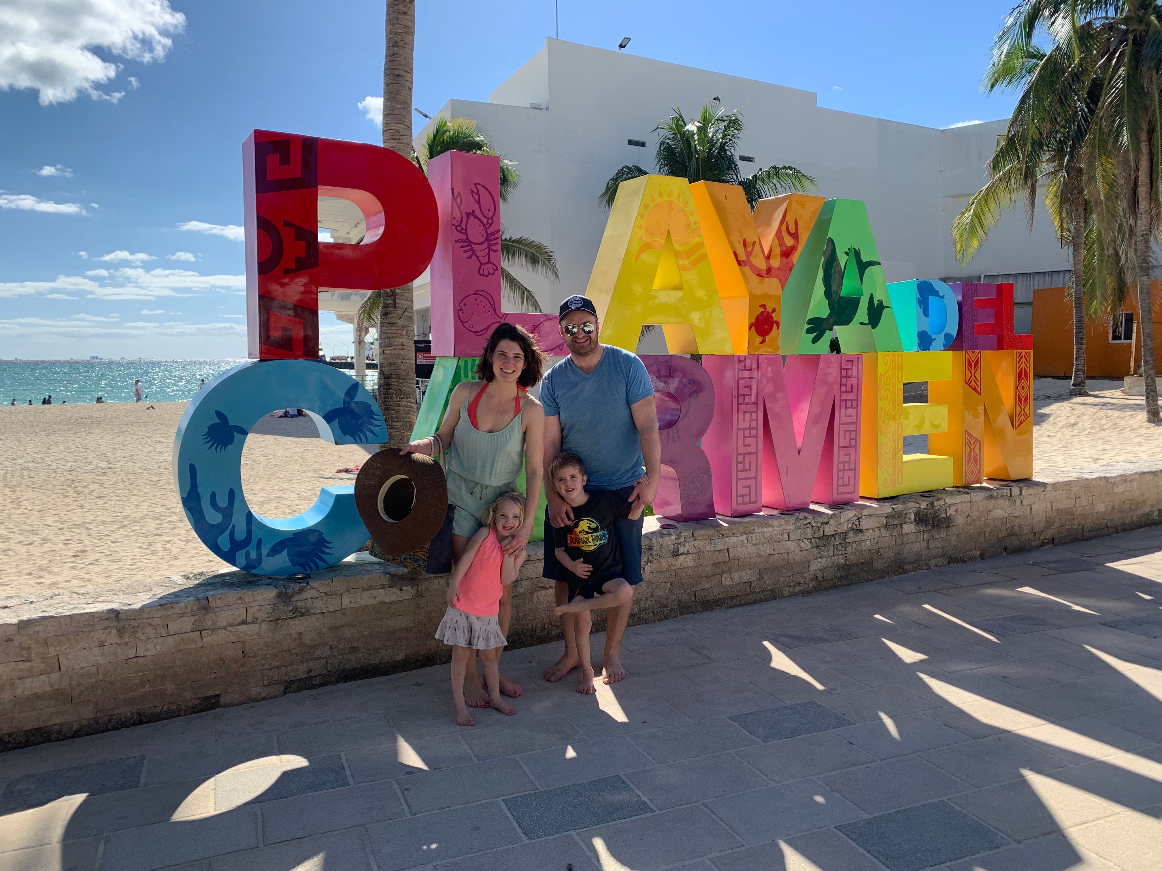 Two adults with two children standing in front of them, all standing in front of a large, colourful sign that reads ‘Playa Del Carmen’ in front of a beach.