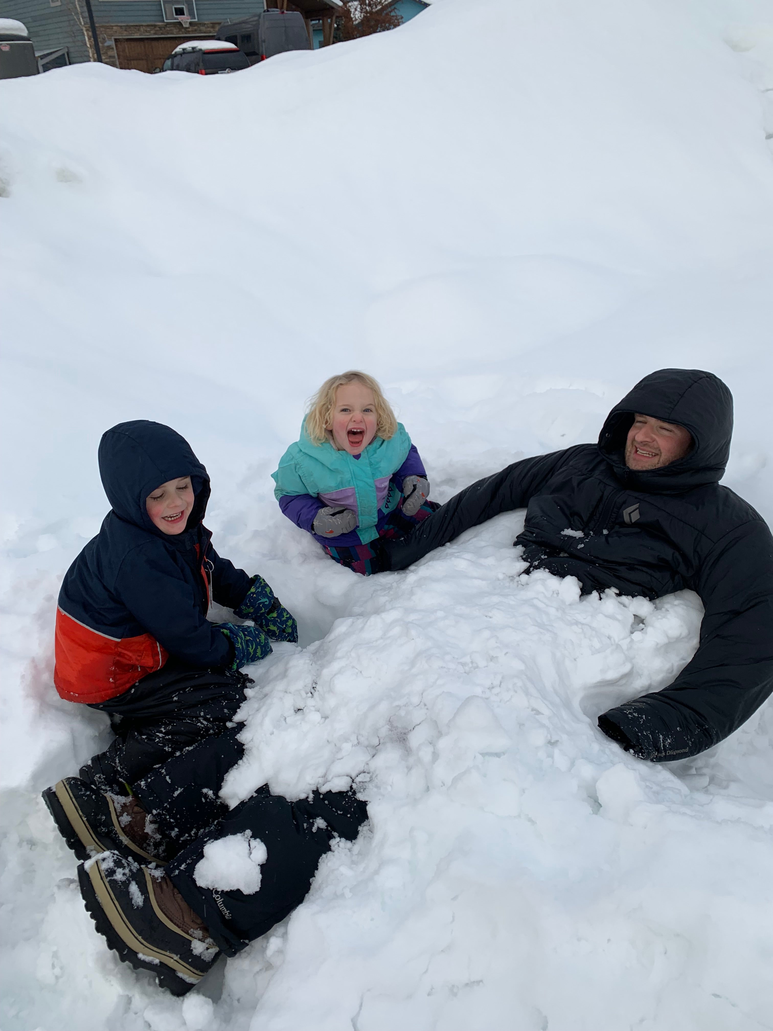 Two children burying an adult in the snow. All are smiling.