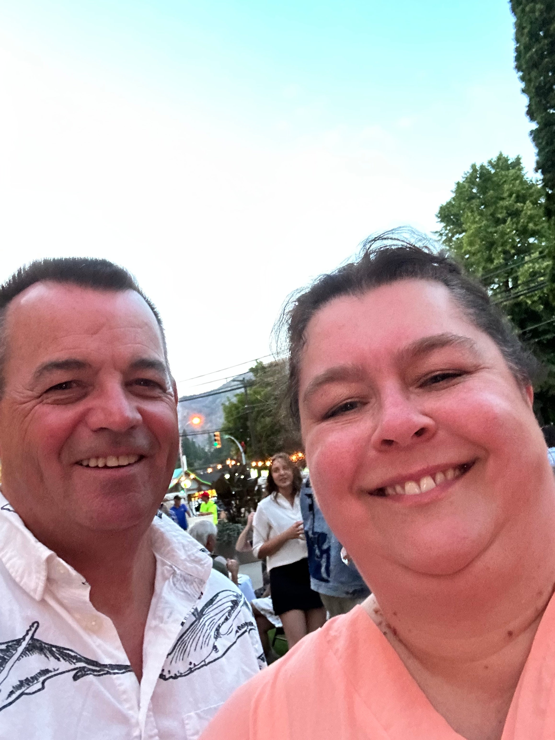 A man and woman take a selfie together outside with a group of people in the background. 