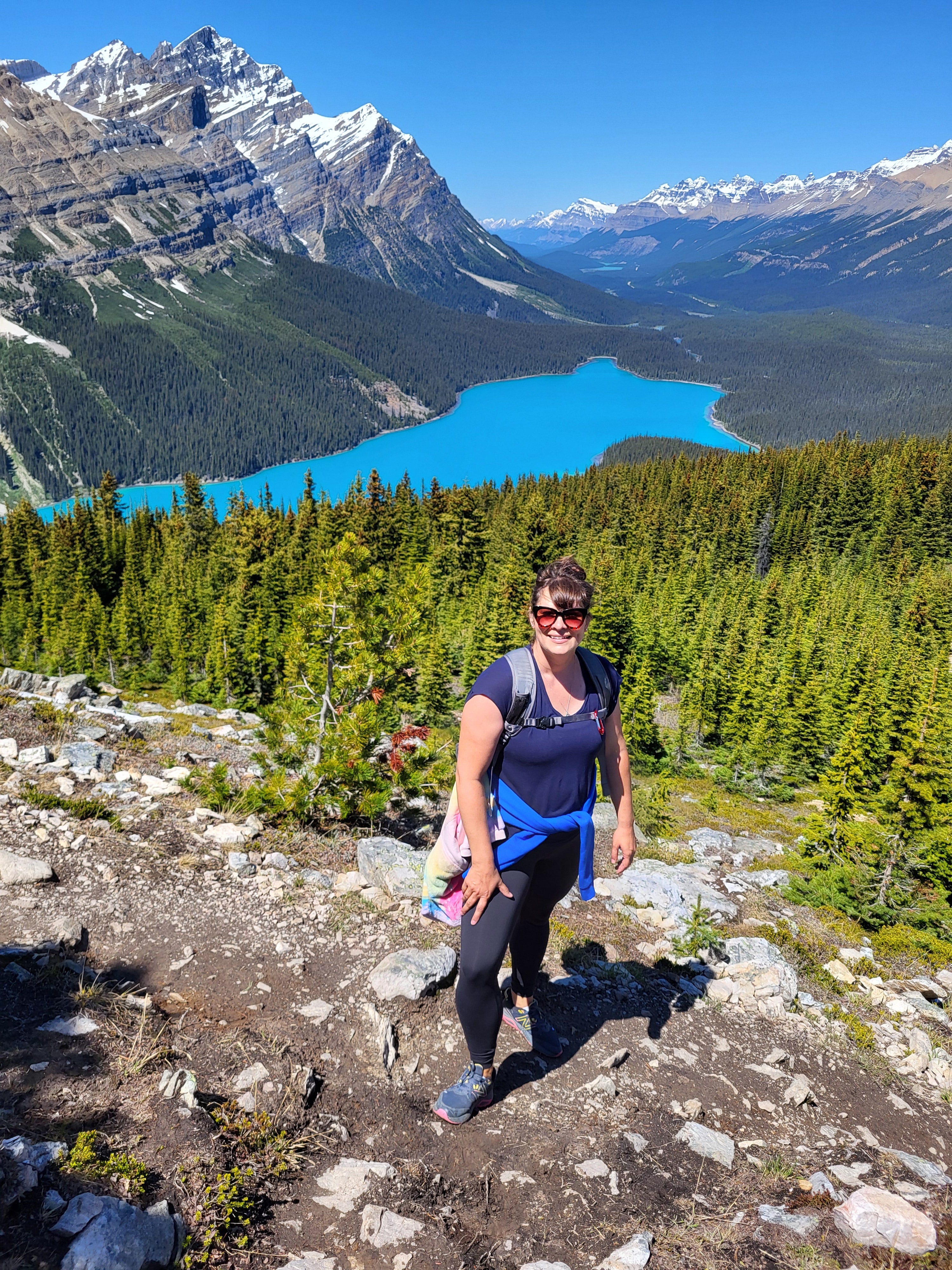 A person wearing a backpack on a hiking trail with a forest, lake and mountain in the background.