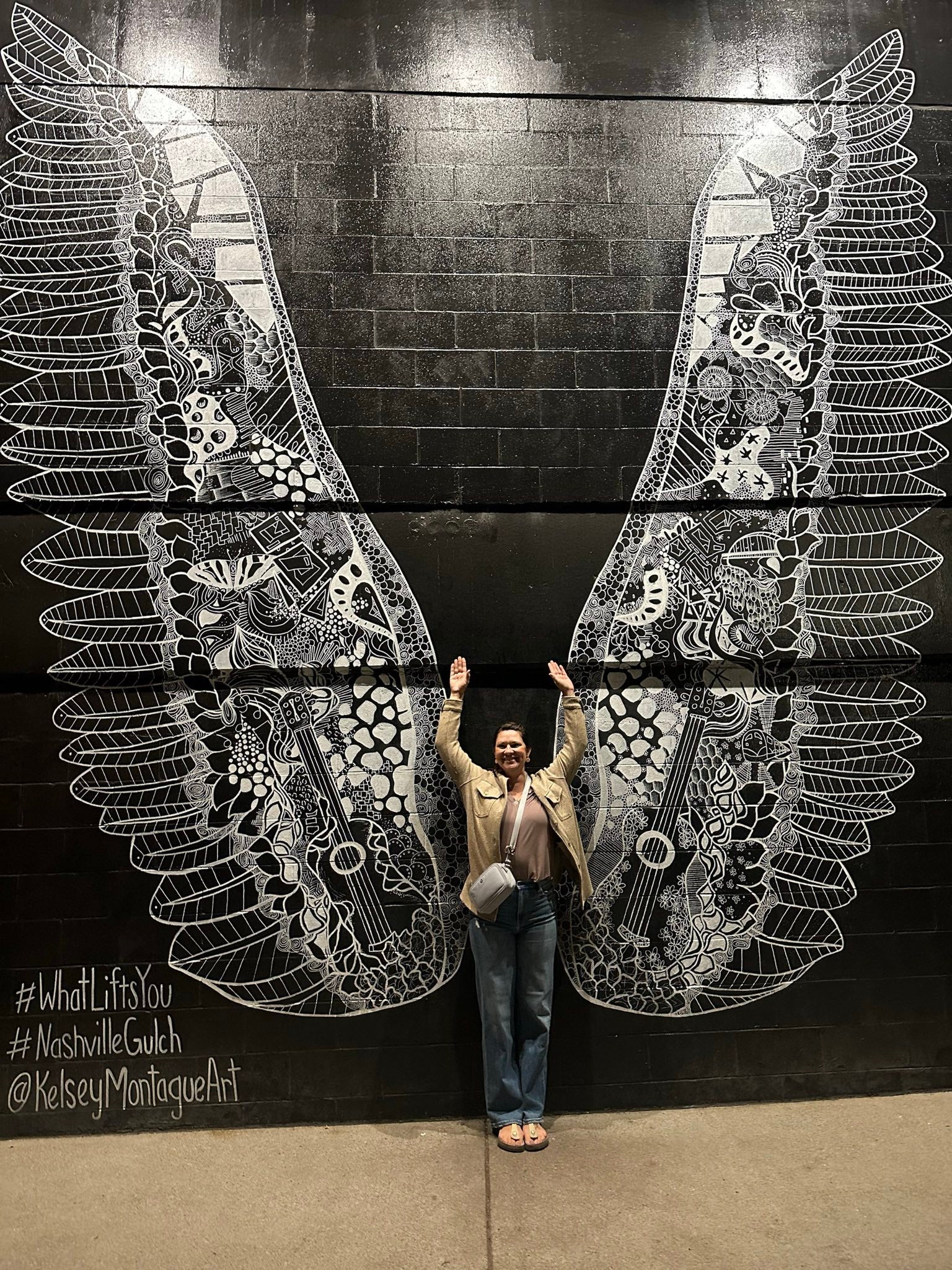 A person standing with outstretched arms in front of a mural of large wings.