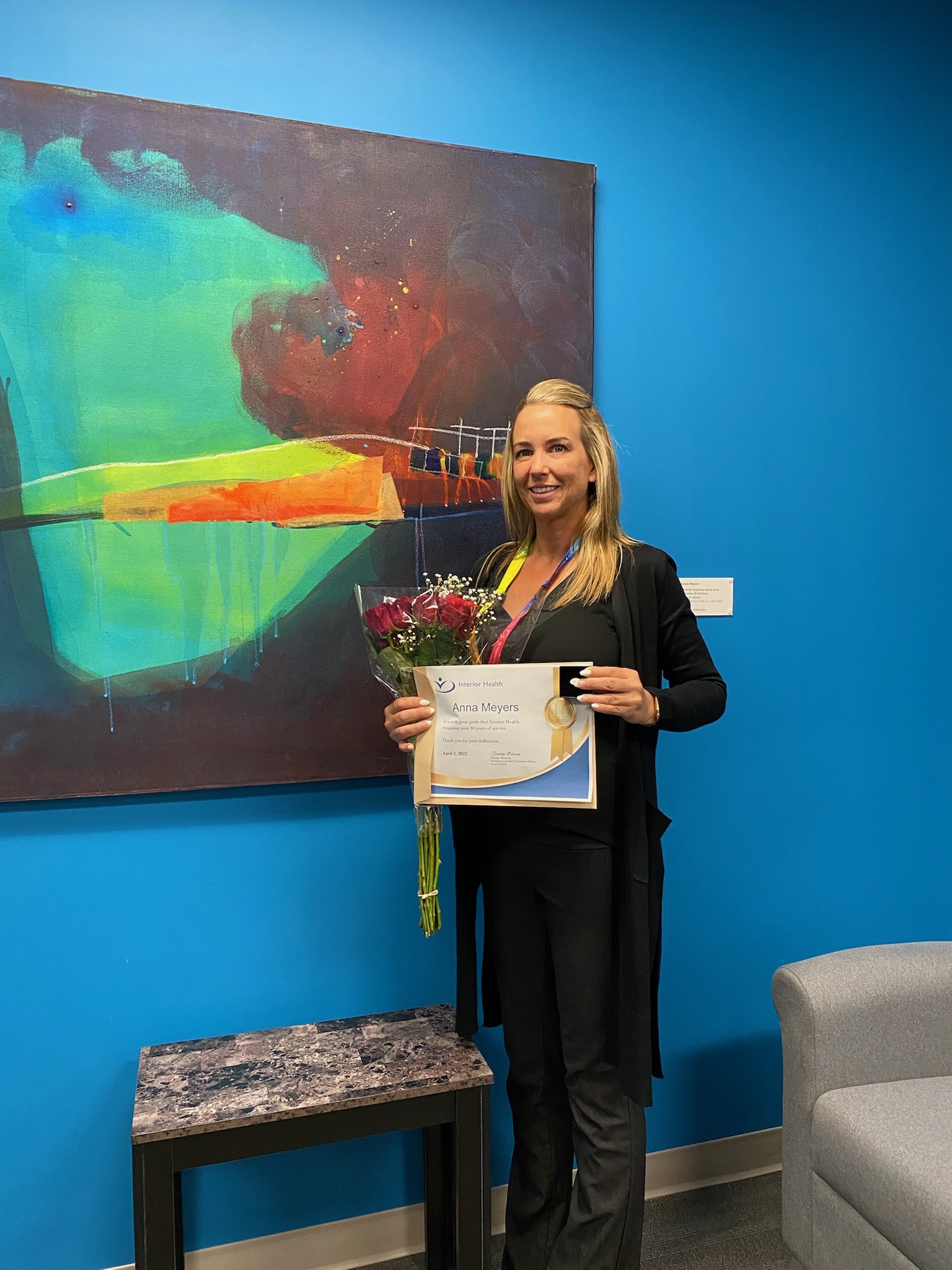 A woman dressed in black standing on front of a blue wall with a piece of art, holding a years of service certificate and a bouquet of roses.