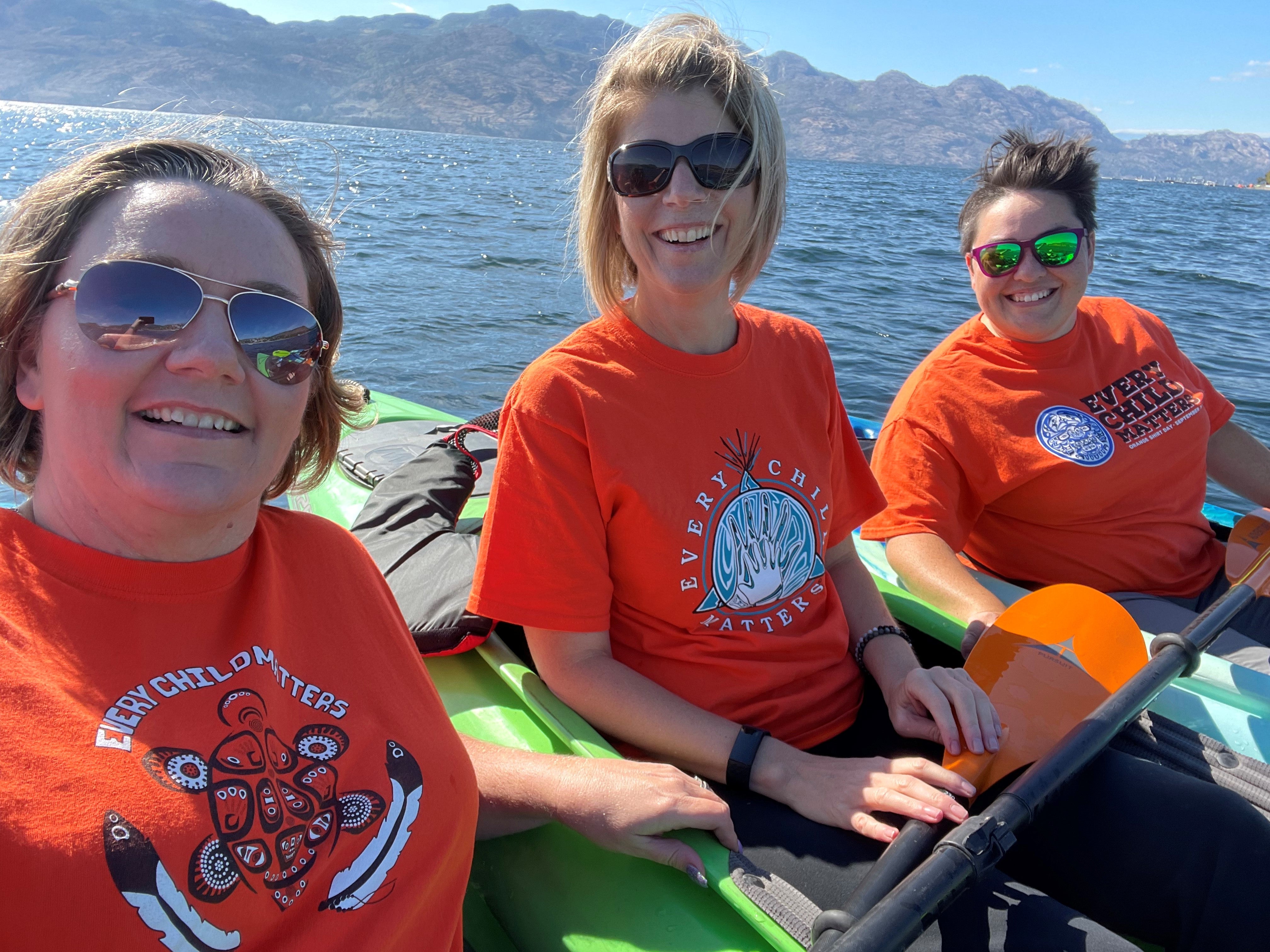 Three people in green kayaks on a lake, wearing orange shirts.