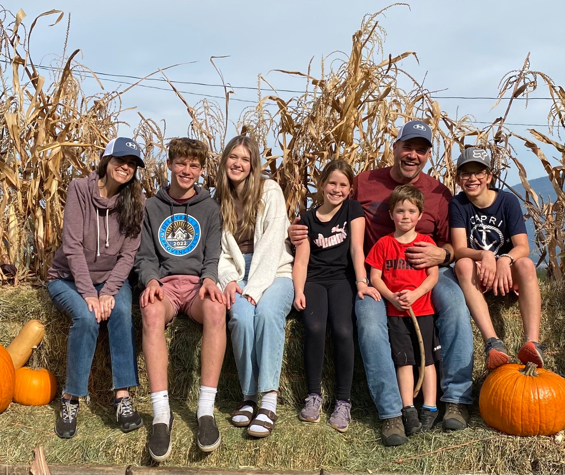 Man, woman, and five kids of various ages sitting next to pumpkins with corn stalks behind them.