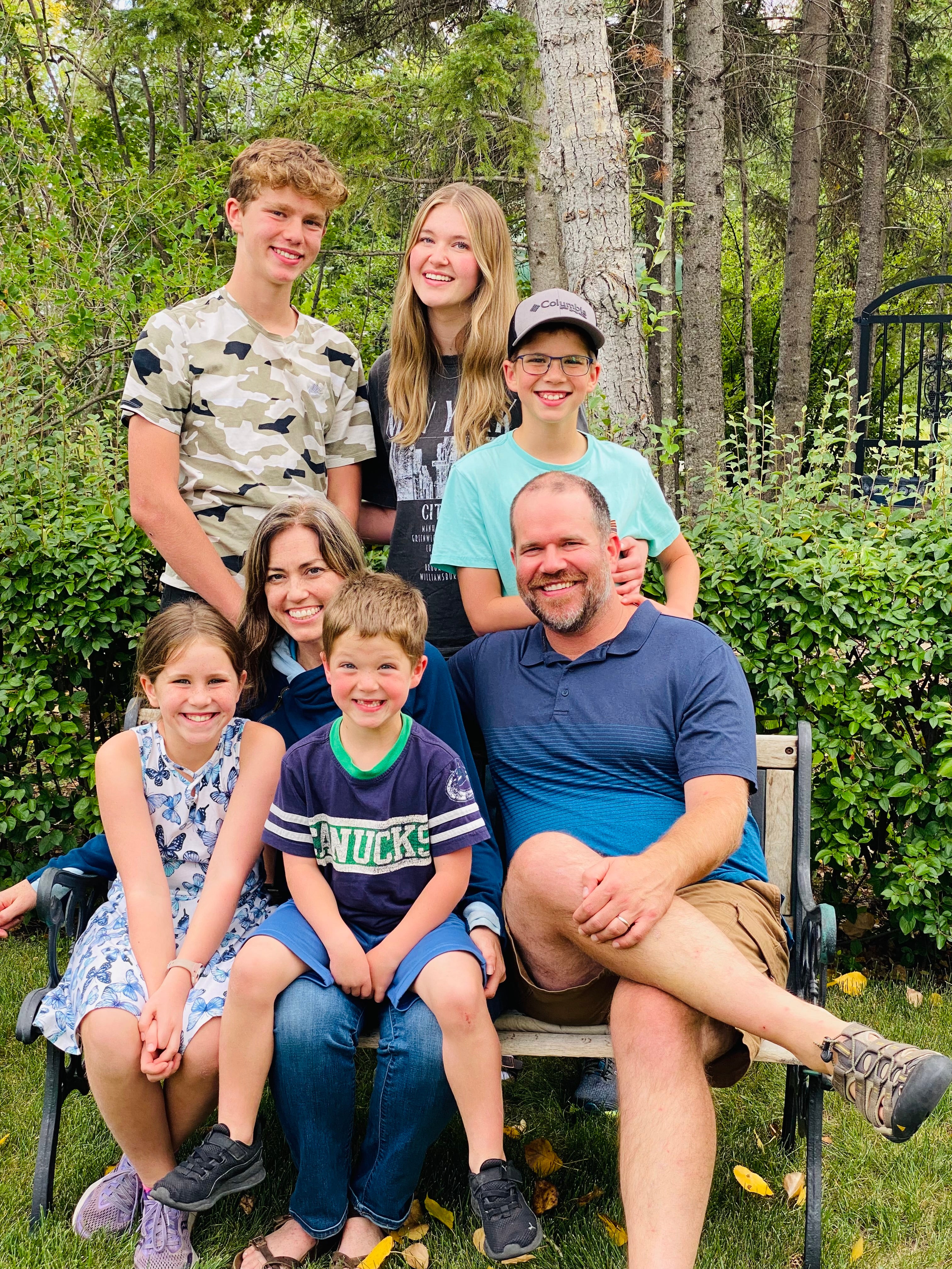 Man, woman, and five kids of various ages sitting on a park bench with trees behind them.