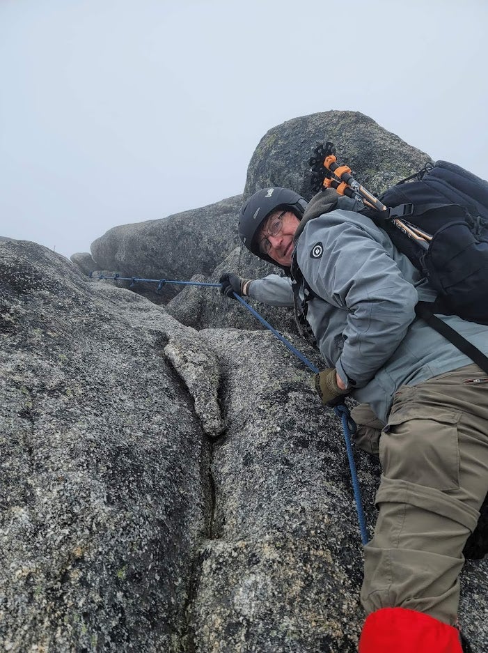 Man wearing a helmet, backpack, grey jacket and khaki pants, looking down from climbing a grey rock face.