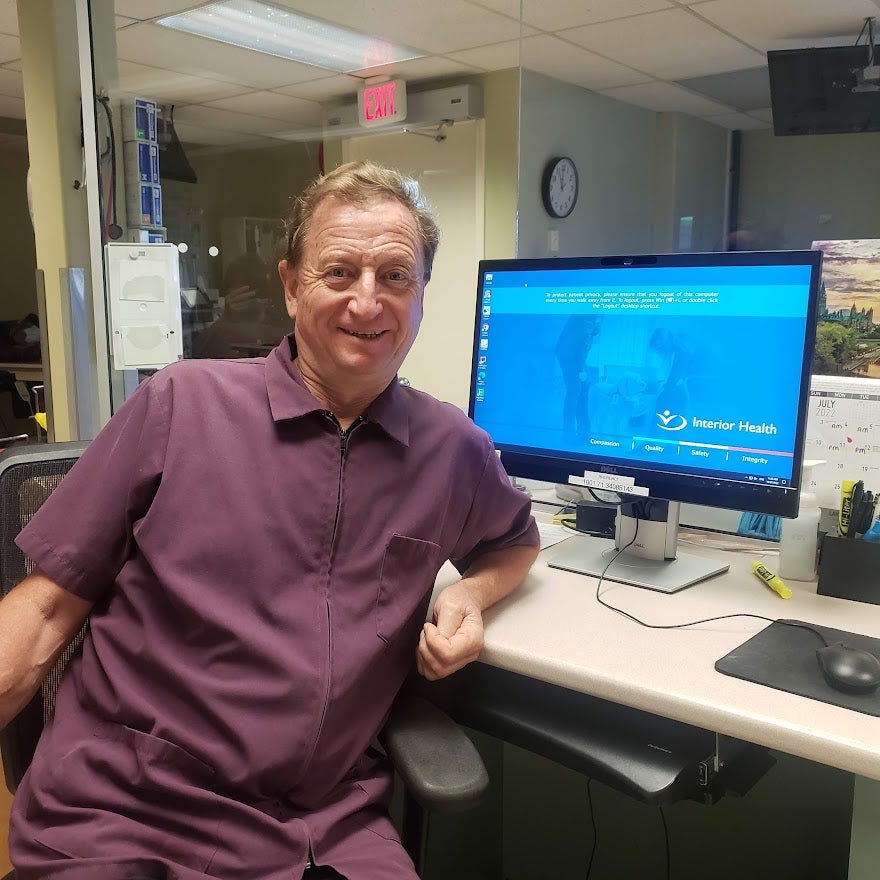 Man wearing a short-sleeved purple shirt, sitting at desk with blue screen on computer monitor.