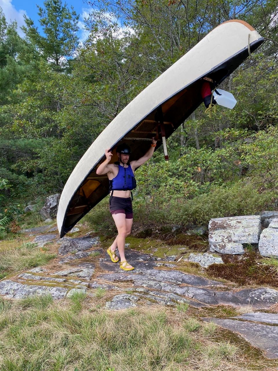 A man in a blue life preserver, maroon swim shorts and yellow sandals carrying a beige canoe with a forest in the background.