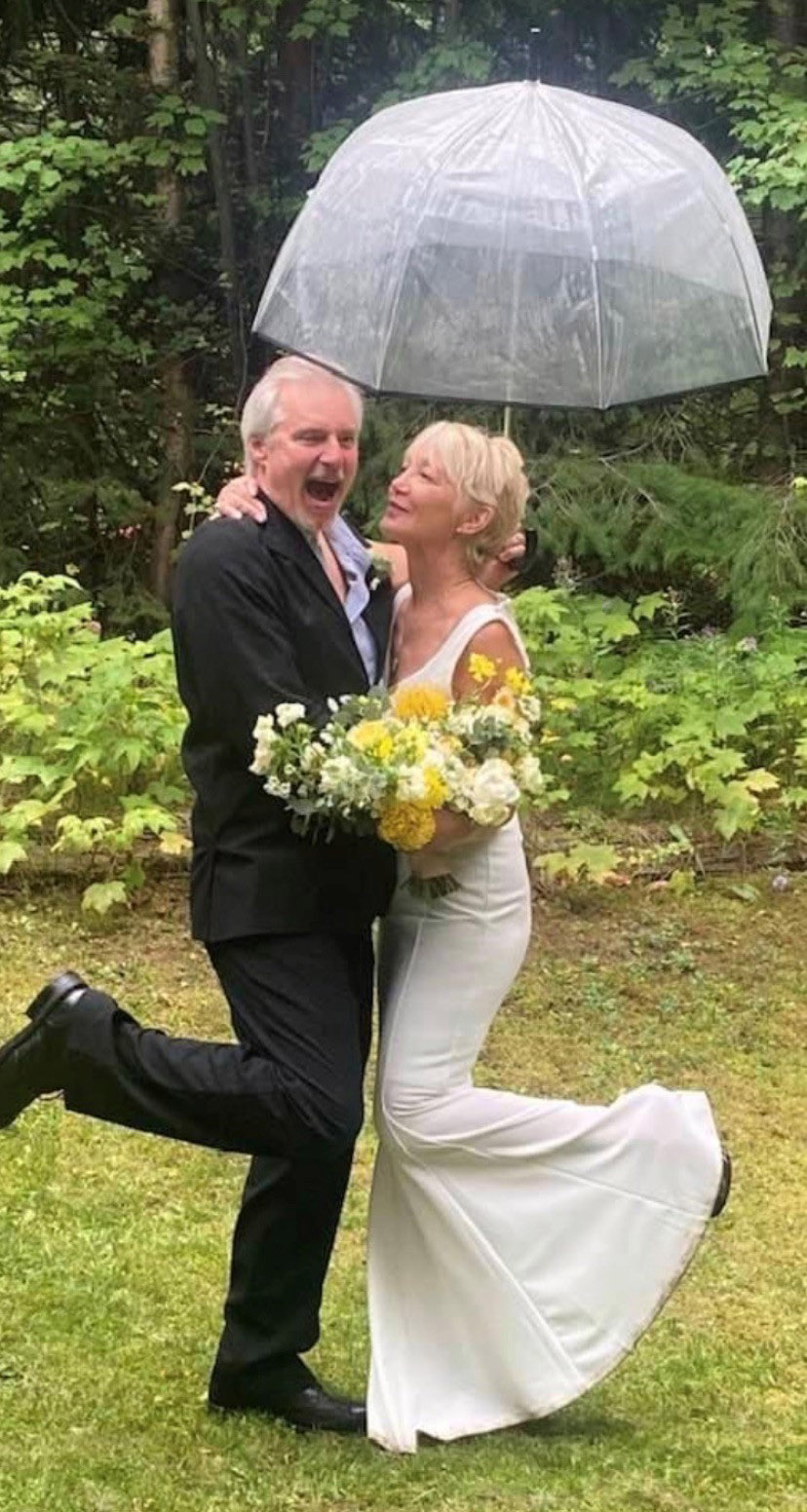A man with grey hair on the left wearing a black suit, with his right leg up while standing on the left, and holding an umbrella, facing a woman with blonde hair in a wedding dress, holding a bouquet of white and yellow flowers, with her left leg up while standing on the right.
