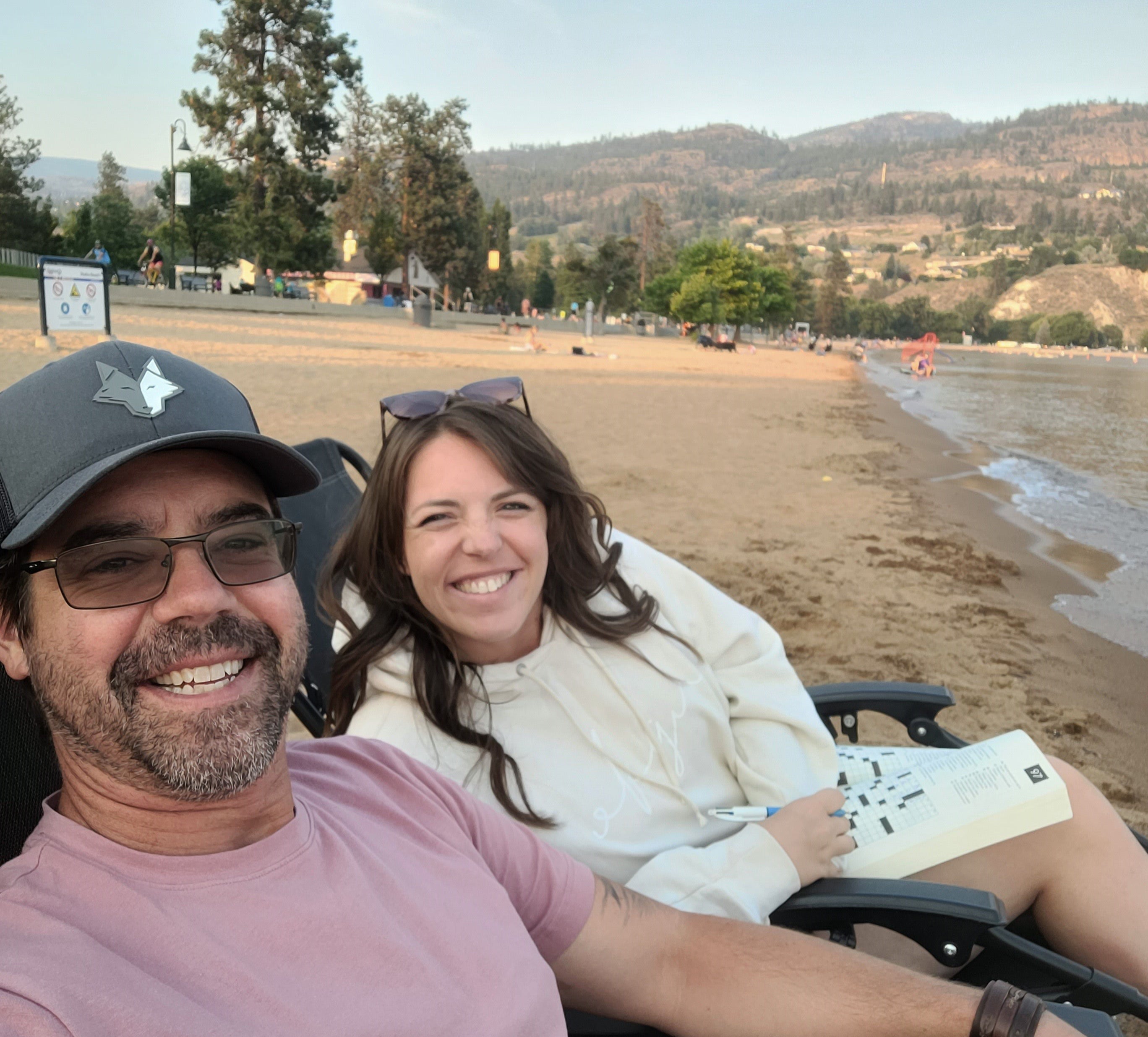 Smiling man with moustache and beard wearing grey ballcap, sunglasses and light pink t-shirt, sits next to smiling woman with long brown hair, wearing sunglasses, and white jacket, sitting with arms folded on lake beach with trees and mountains in the background
