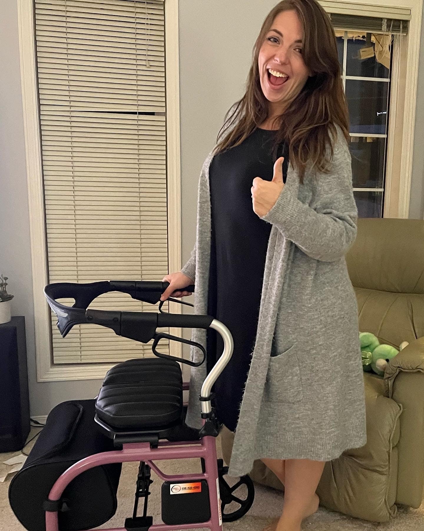 Smiling woman with long brown hair wears a black dress and long grey sweater and gives a thumbs up standing behind a walker, a long narrow window with horizontal blinds and recliner chair in the background