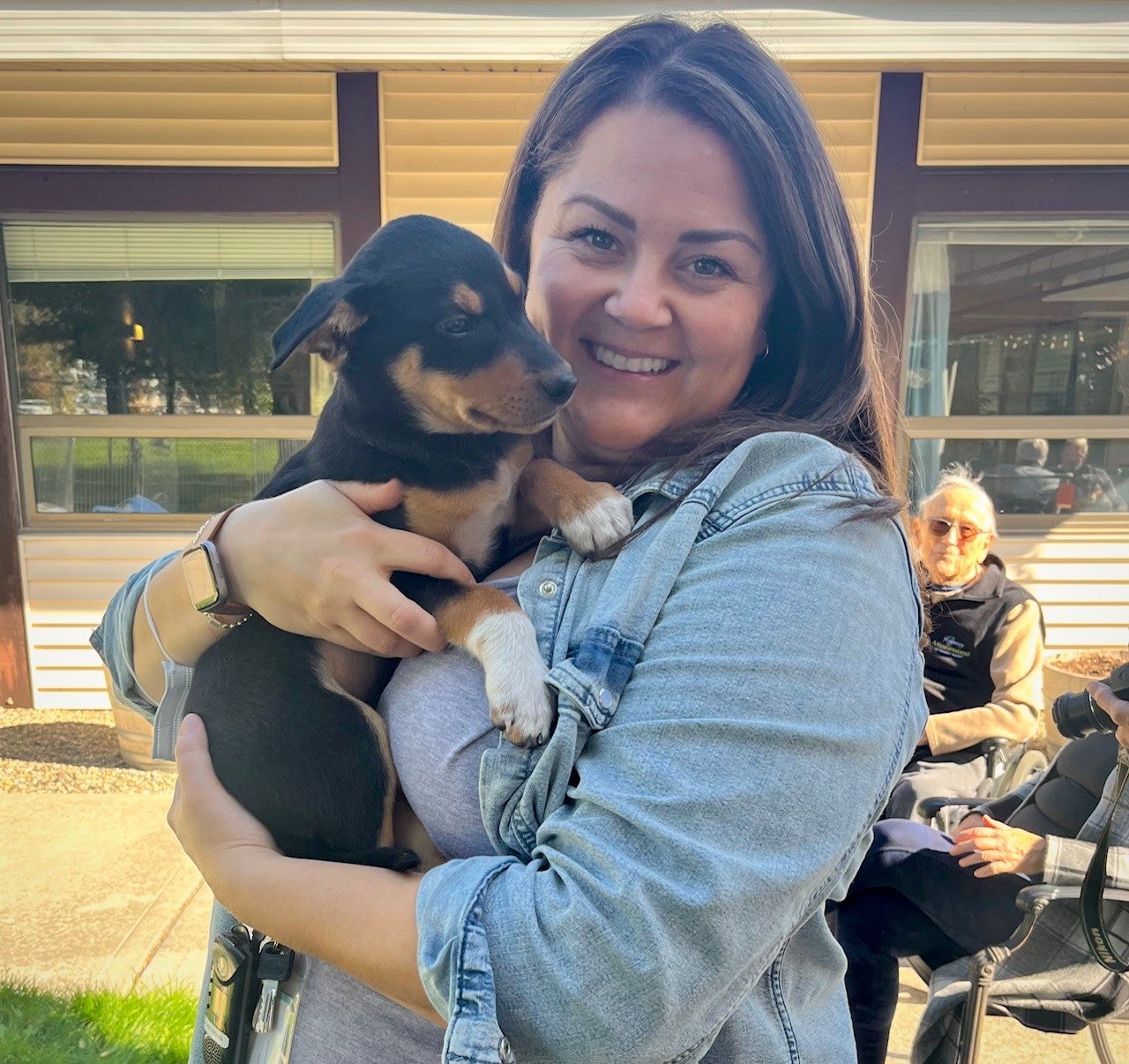 A woman with long brown hair wearing a jean shirt, holding a small black and brown dog, poses for a picture 