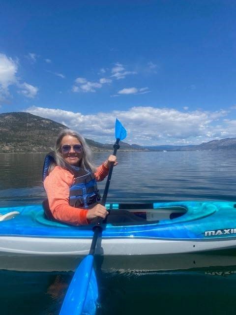 A woman with light grey hair and sunglasses is sitting in a kayak on a lake holding a paddle 
