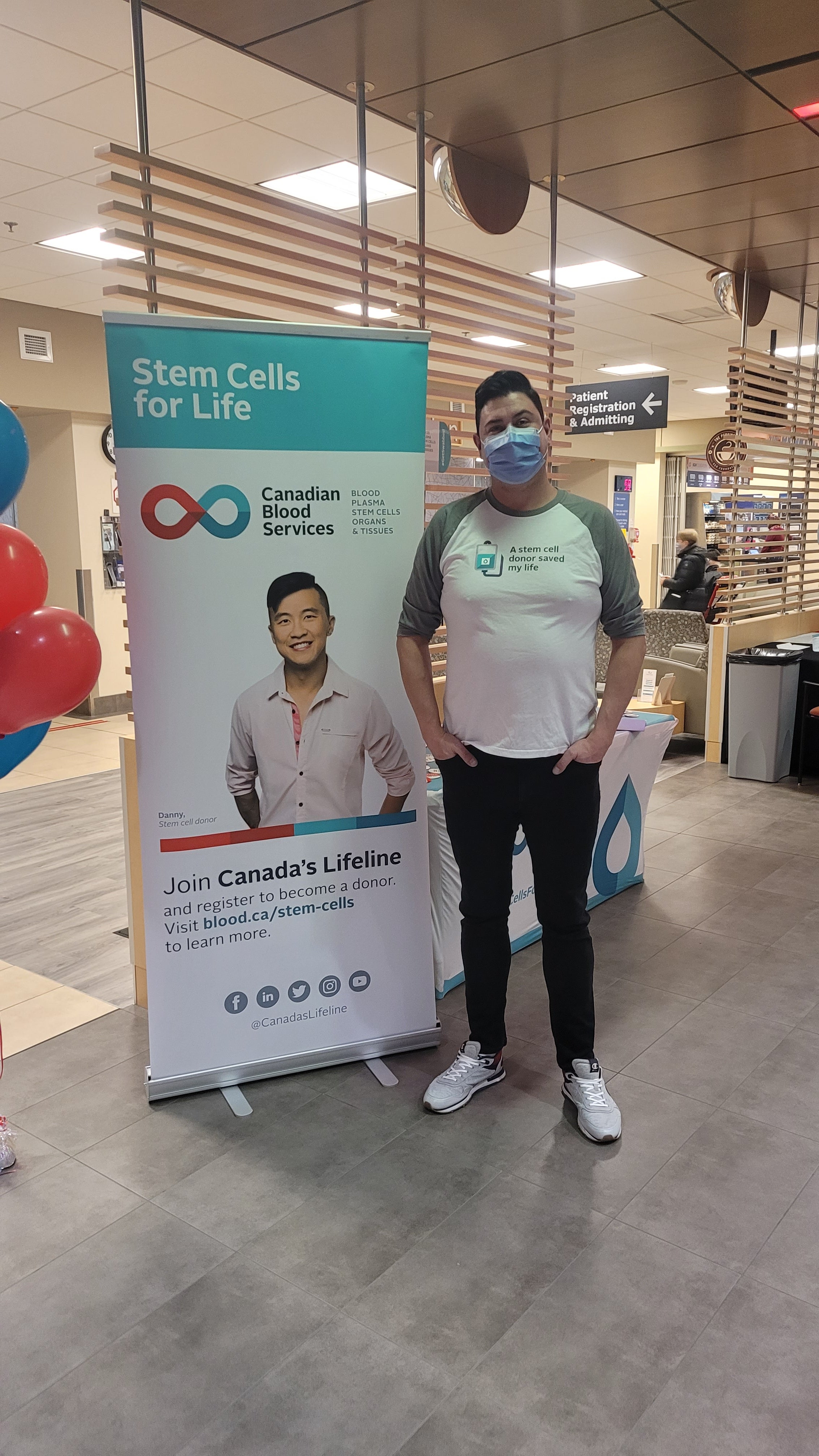 A man standing in a hospital lobby with his hands in his pockets wearing a face mask, white t-shirt with grey sleeves that says “A stem cell donor saved my life” next to a sign that says “Stem Cells for Life” with an image of a smiling man in a white dress shirt.
