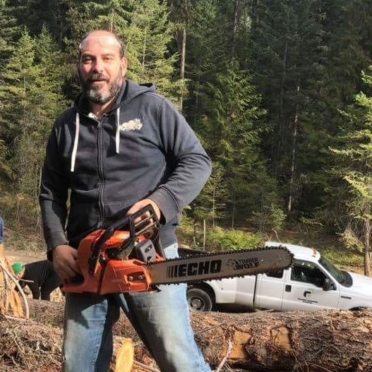 Man in forest holding chainsaw with white truck behind him.