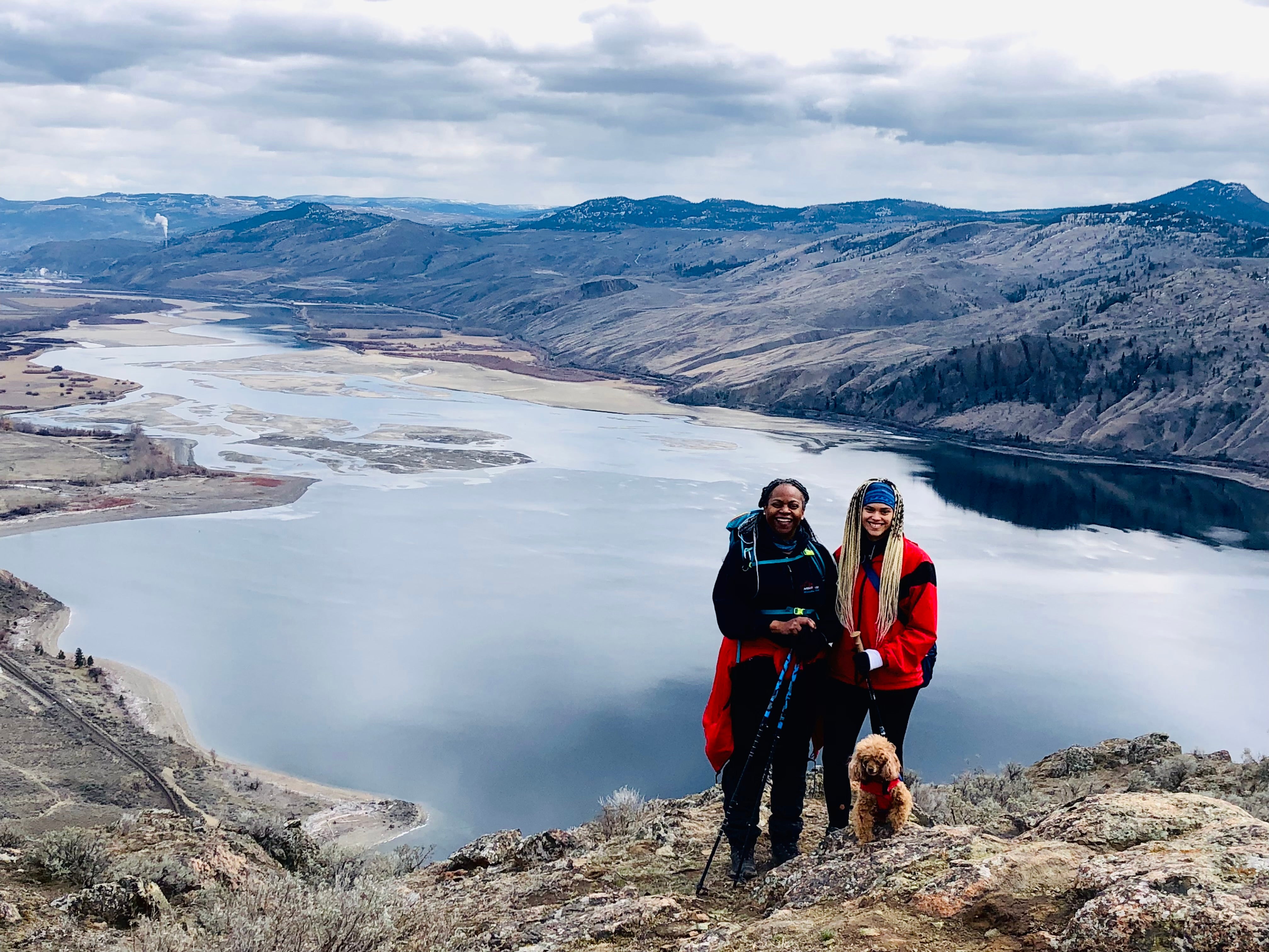 A woman in black and a woman in red at the top of a mountain overlooking a lake.