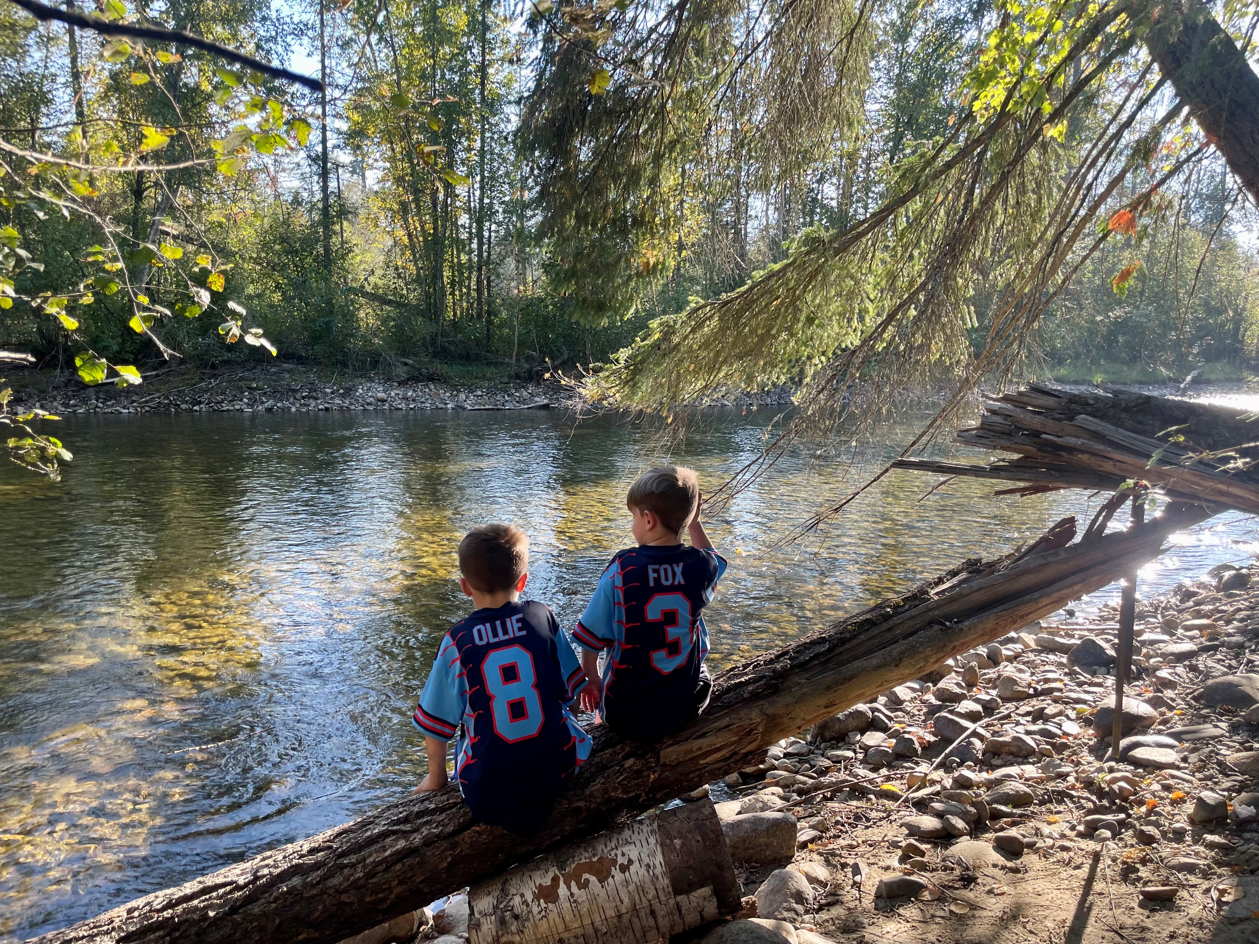 Two boys sitting on a log by a river surrounded by trees.