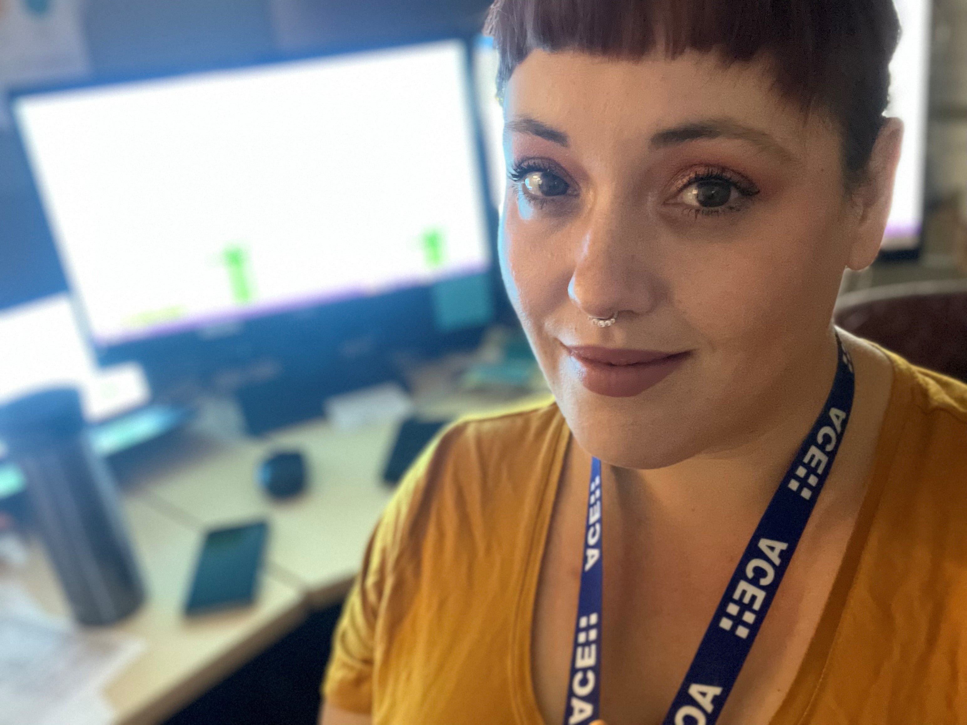 Woman with short brown hair, nose piercing and ACE lanyard has a computer monitor and desk behind her.