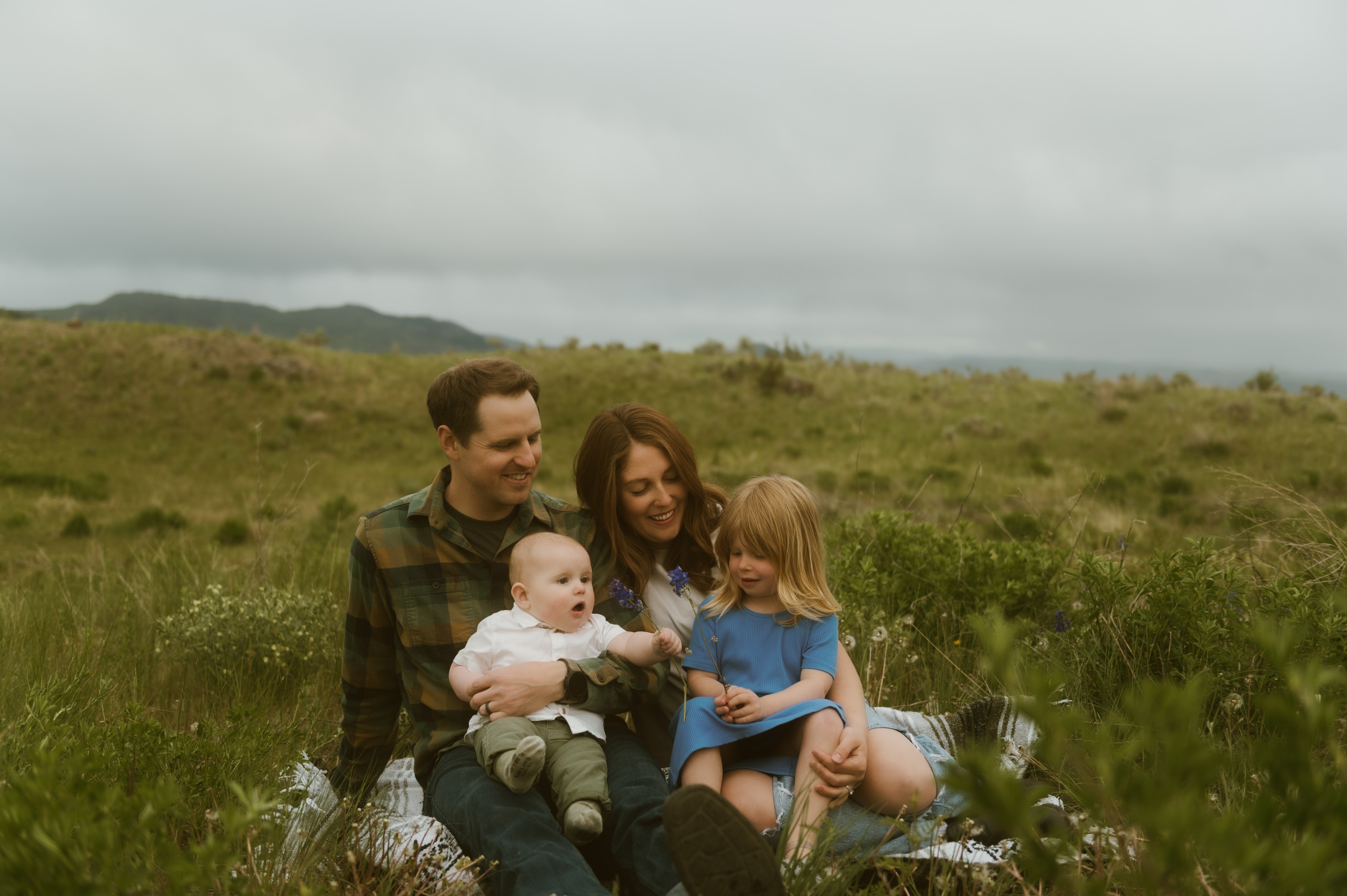 A family of four sitting on a blanket in the grass under an overcast sky.