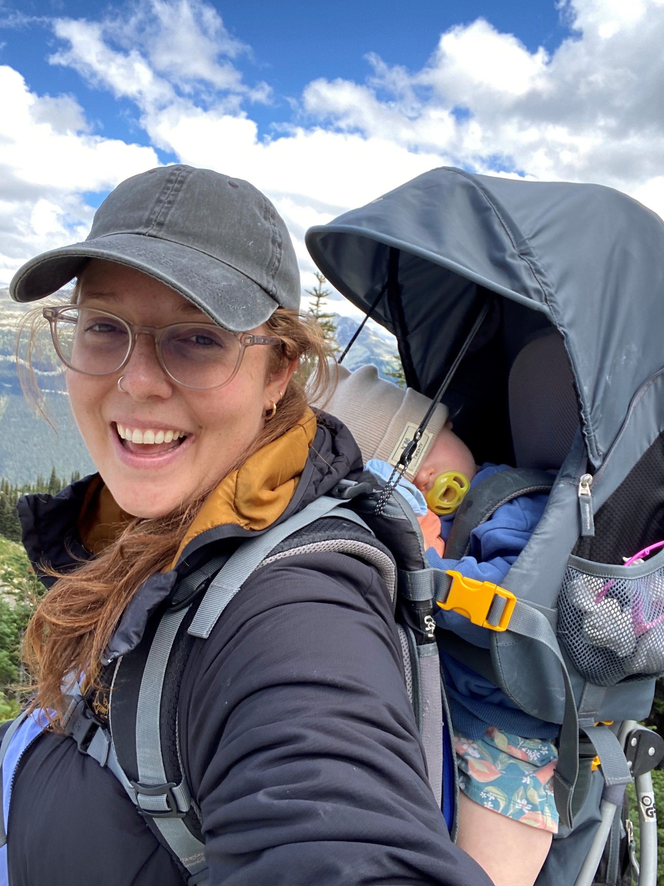 A person in a dark cap and glasses carrying a sleeping child while out on a hike.