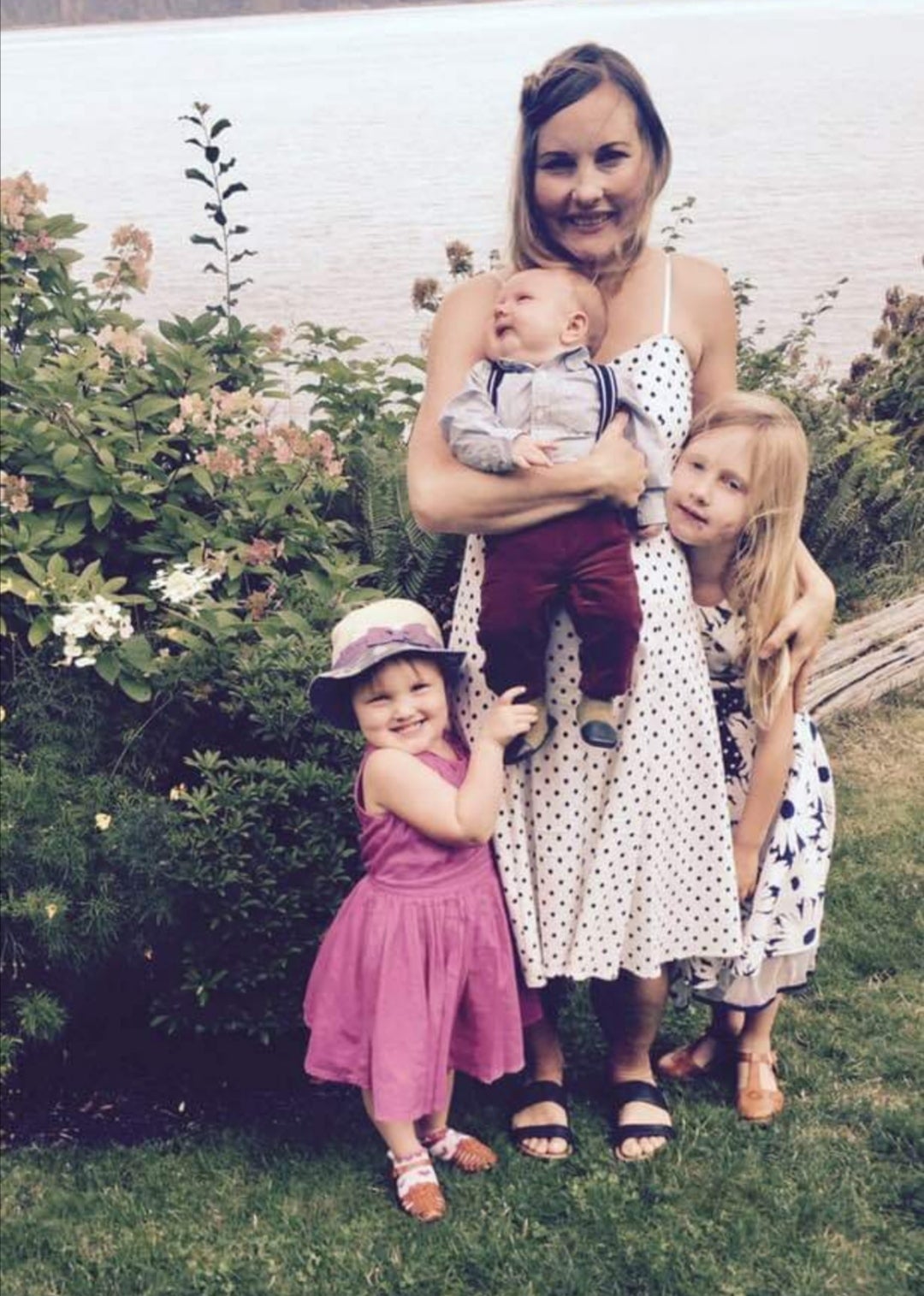 A woman with three children stands together in front of a bush of leaves and flowers and a lake. The woman holds a small baby, and has her arm around a young girl. A toddler smiles and holds on to the baby’s foot. 